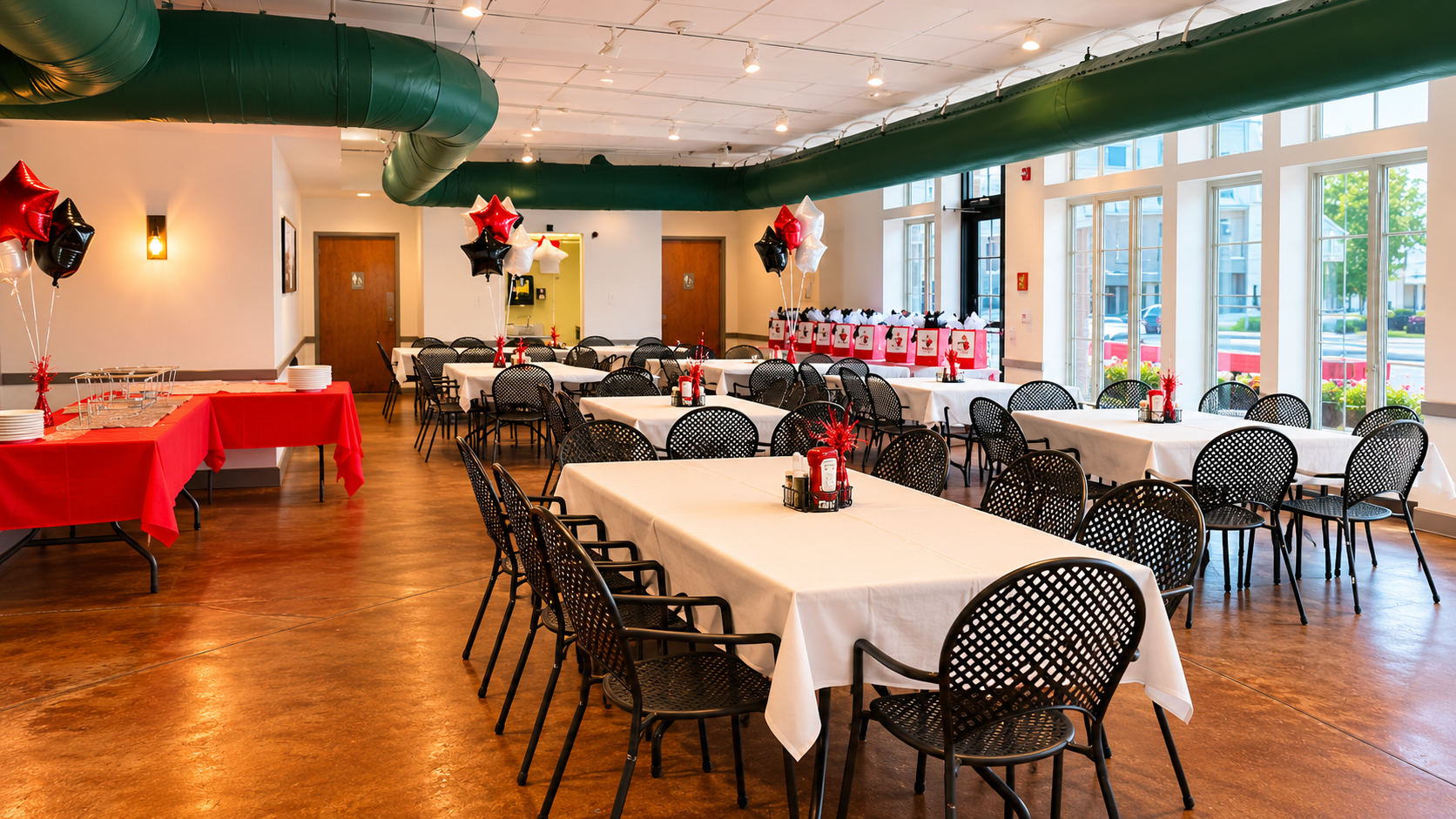 A decorated party room with tables covered in white tablecloths, black chairs, and red, black, and white balloons. There are red gift bags lined up against the windows and a table with red tablecloths holding plates and cups, with some red décor.