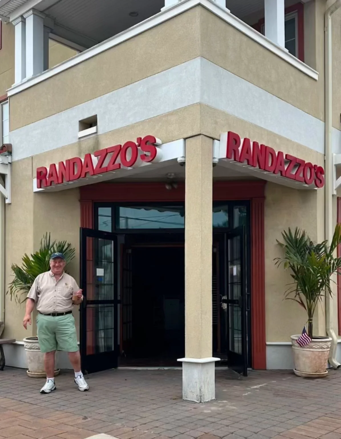 A man standing outside the entrance of Randazzo's restaurant, smiling and giving a thumbs-up. The restaurant has a beige exterior with red signage and two potted plants near the entrance.
