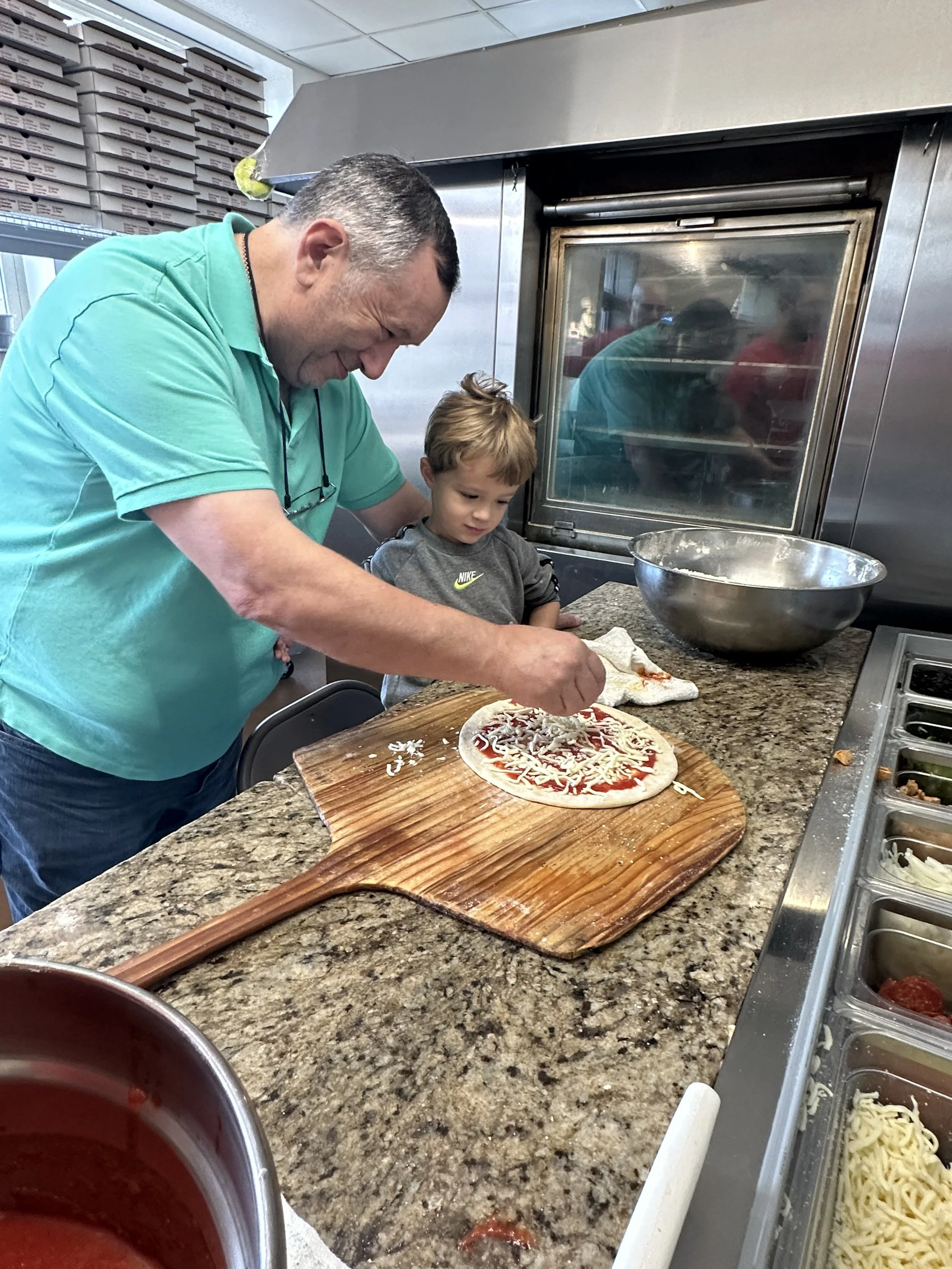 A man and a young boy making pizza together in a commercial kitchen. The man is sprinkling cheese on the pizza while the boy observes, standing behind a wooden pizza peel on a granite countertop.