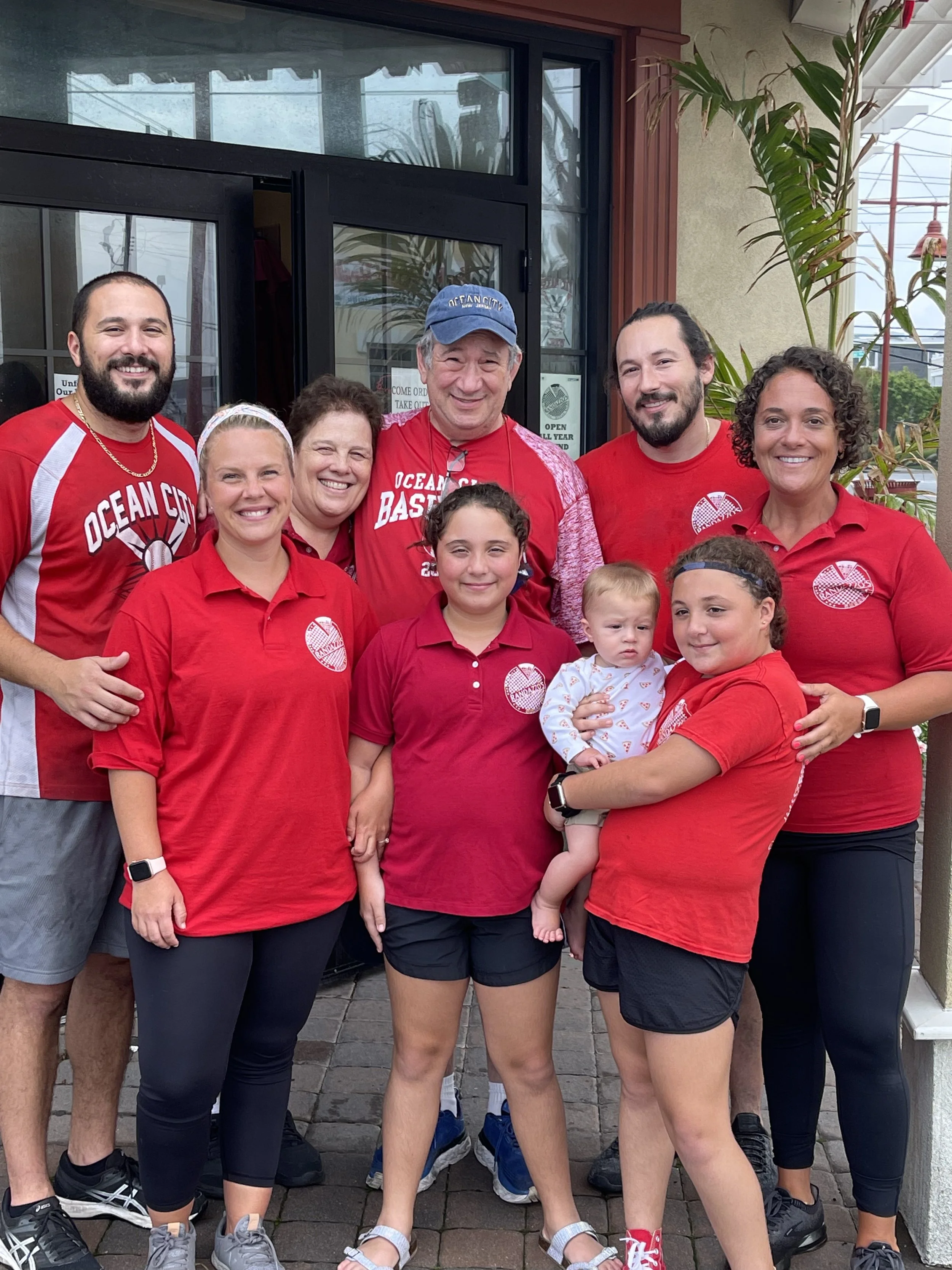A group of people standing together outside a storefront, smiling for the camera, wearing red shirts with a logo, some children in front, some adults and children with diverse appearances, some with curly hair, some with beards, in front of a glass door and window surrounded by plants and a brick wall.
