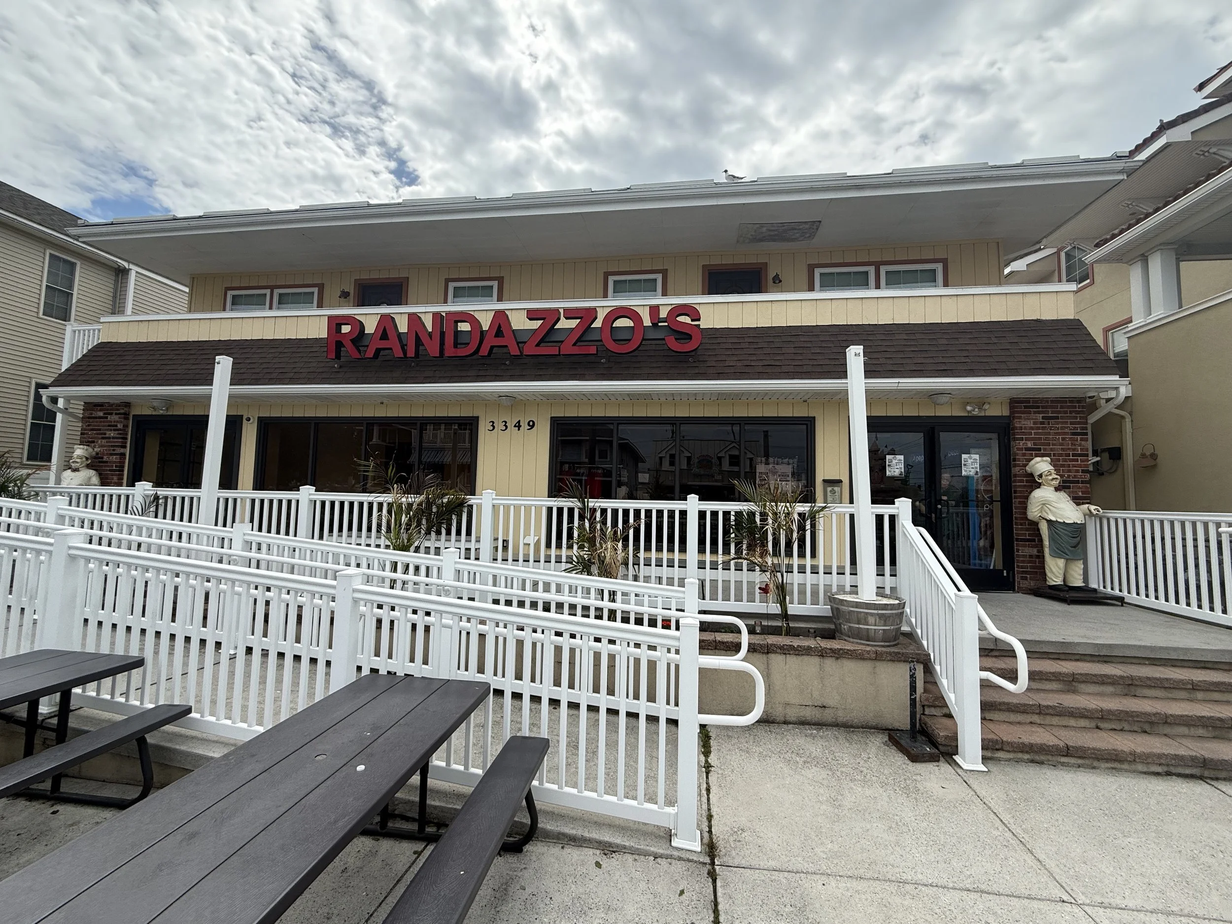 Exterior of a restaurant called Randazzo's with outdoor seating, a white fence, and a large sign on a beige building. There are statues of chefs and a cloudy sky above.
