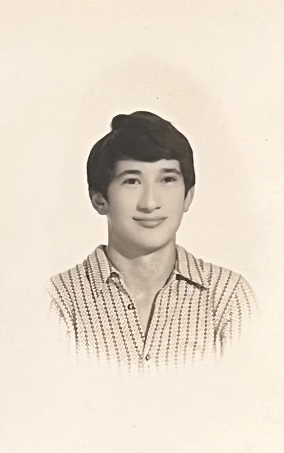 A black-and-white portrait of a young man with short, dark hair, wearing a patterned collared shirt, smiling slightly against a plain background.