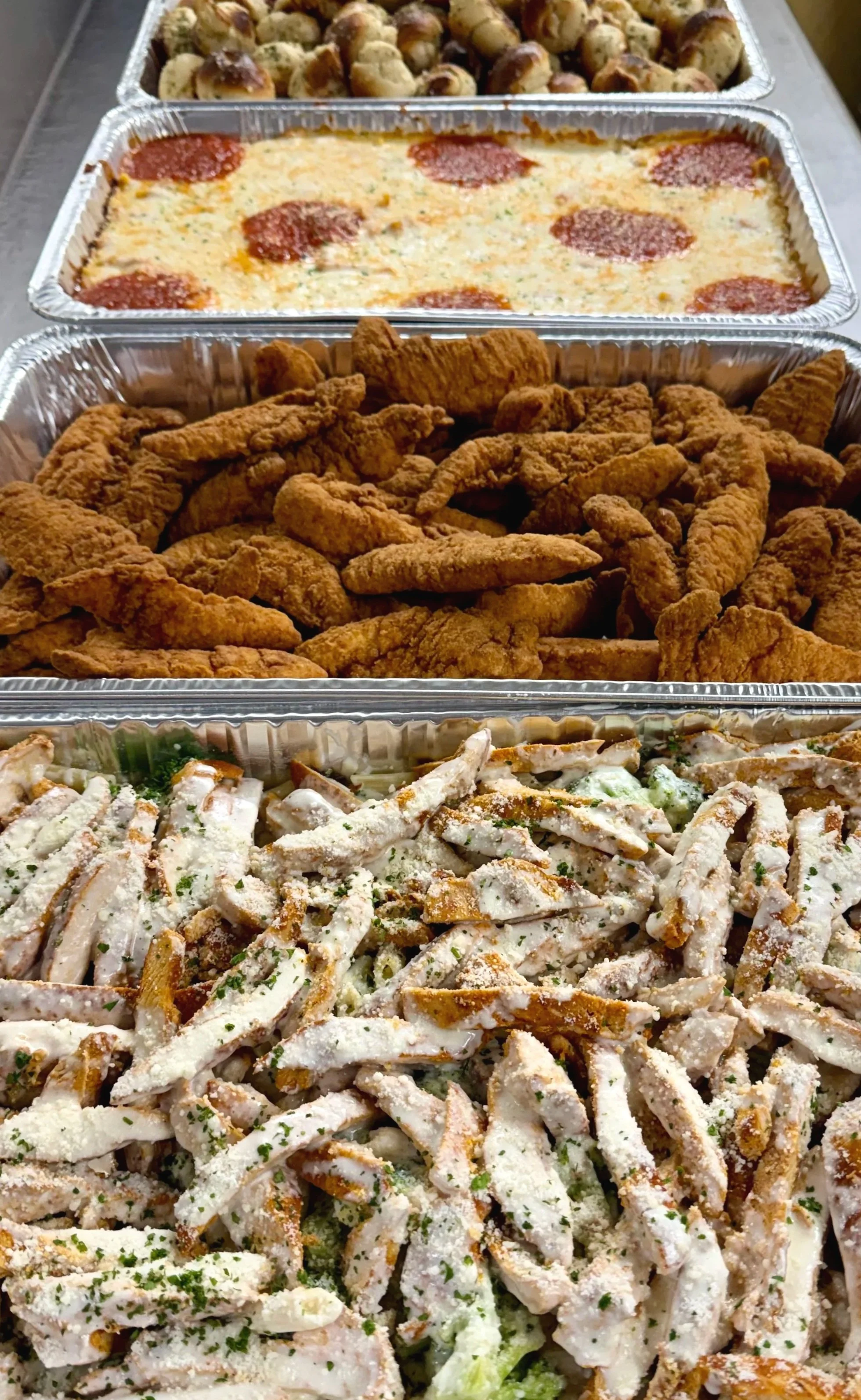 Three aluminum trays with food: top tray with small baked potatoes, middle tray with baked lasagna, bottom tray with chicken Caesar salad.