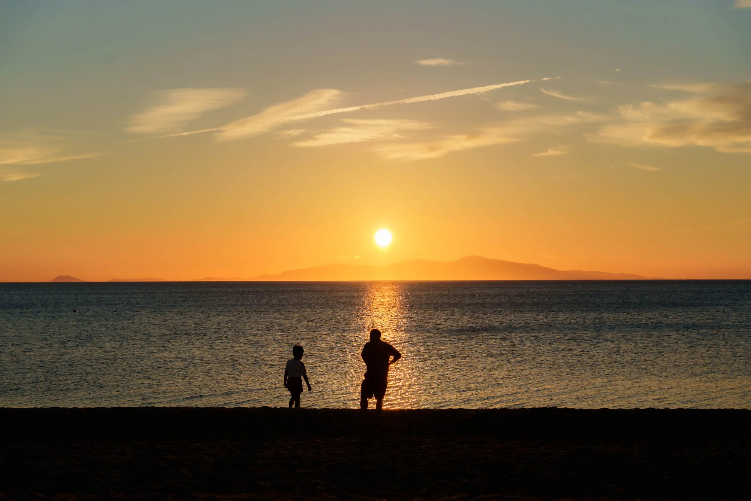 夕日が沈む海と空、山々のシルエットと海辺で遊ぶ子供と大人のシルエットの風景