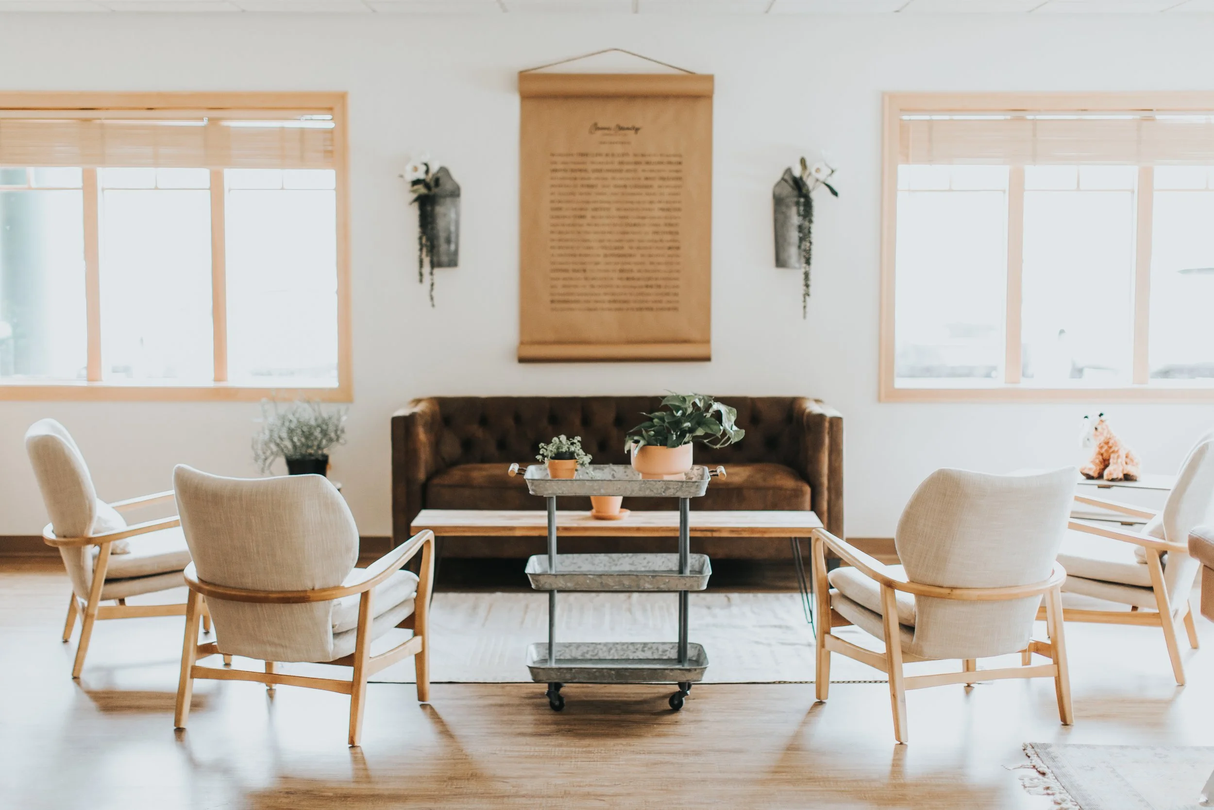 A bright and cozy living room or sitting area with a beige and white color scheme. Central features include a dark brown tufted sofa, surrounded by four white armchairs with wooden frames, and a marble top cart with plants. The walls are decorated wi