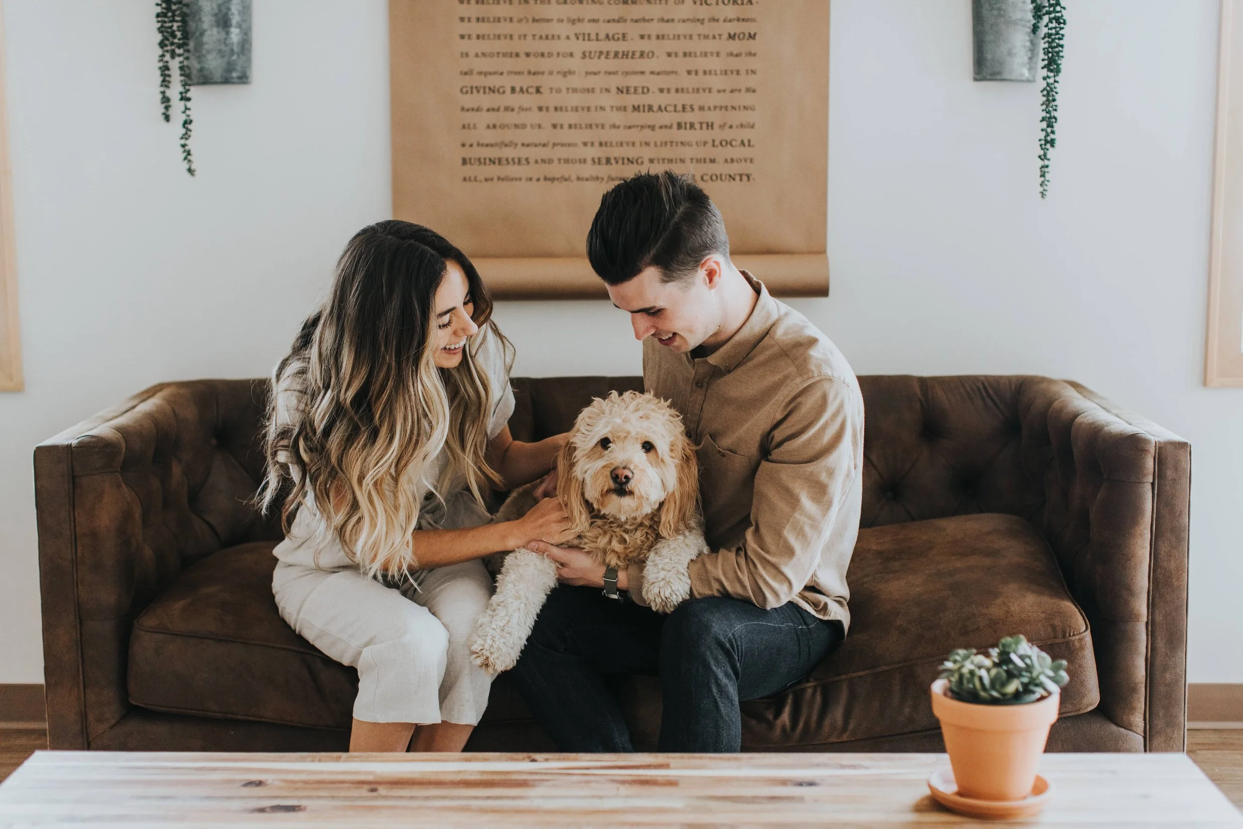 A smiling couple sitting on a brown leather couch, holding and playing with a fluffy light-colored dog in a cozy living room. A brown wooden coffee table with a potted succulent plant is in the foreground. Behind them, there are framed art pieces, gr