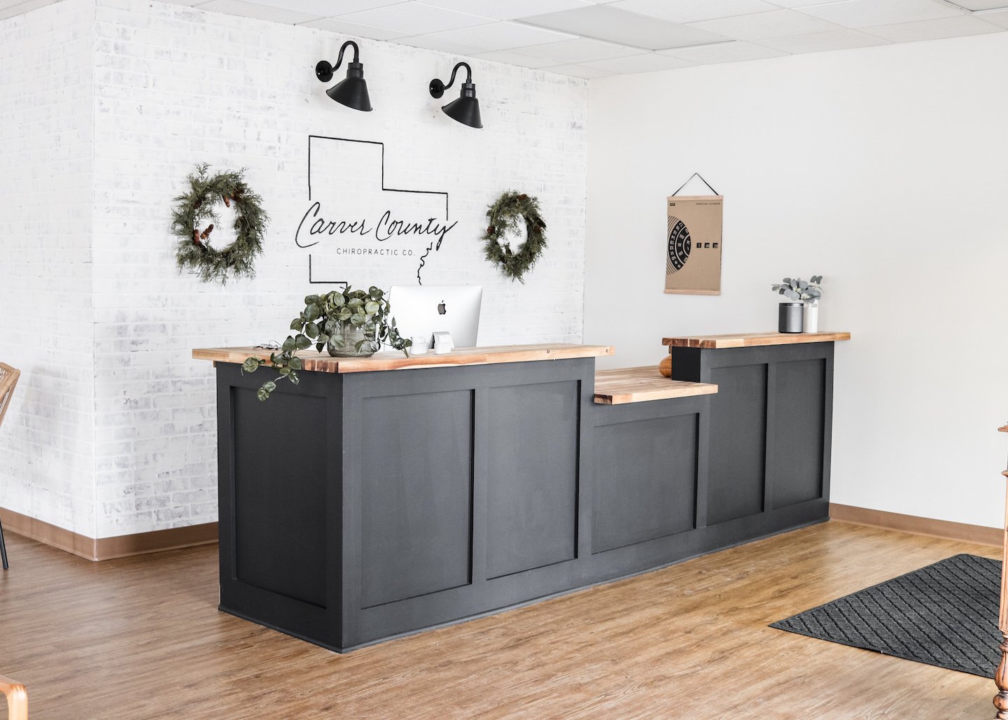 Reception desk at Carver County Chiropractic with white brick wall, black wall-mounted lamps, two Christmas wreaths, and potted plants on desk.