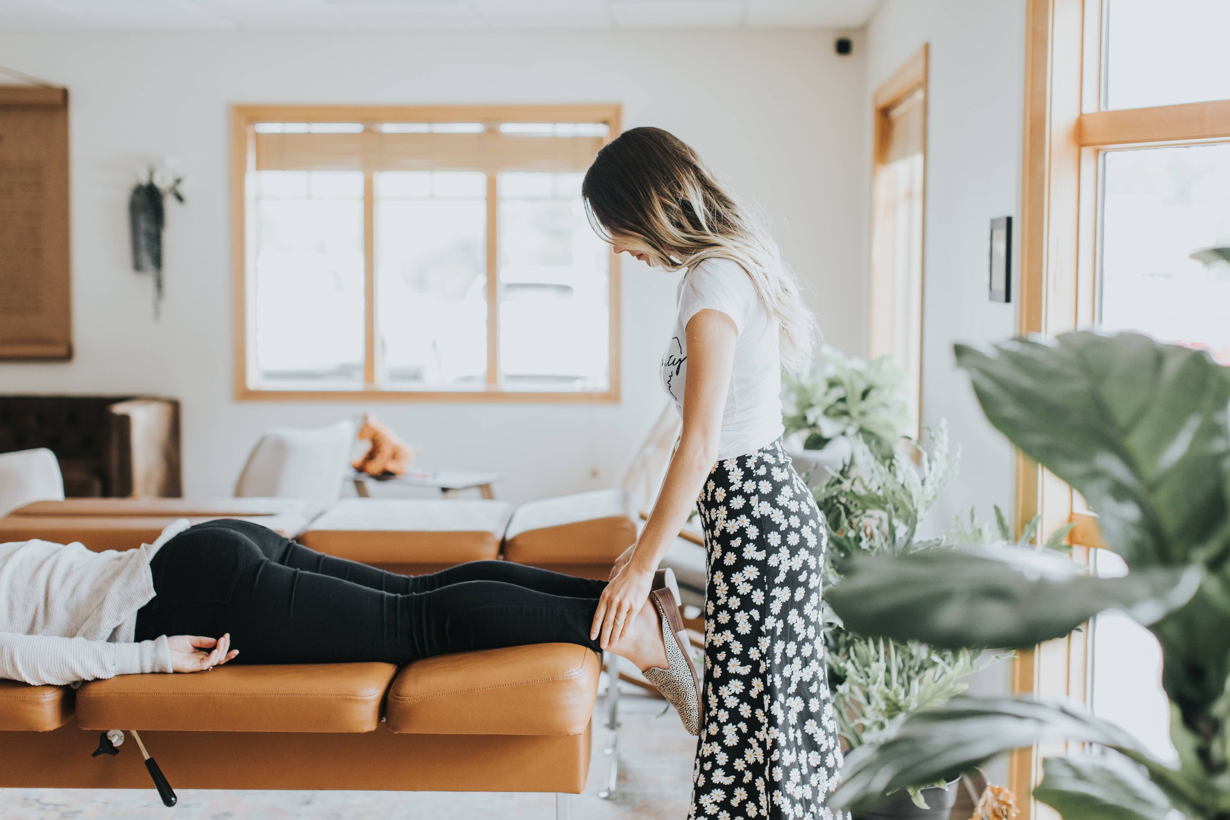 A woman performing a massage on another person lying face down on a massage table in a bright room with large windows and plants.