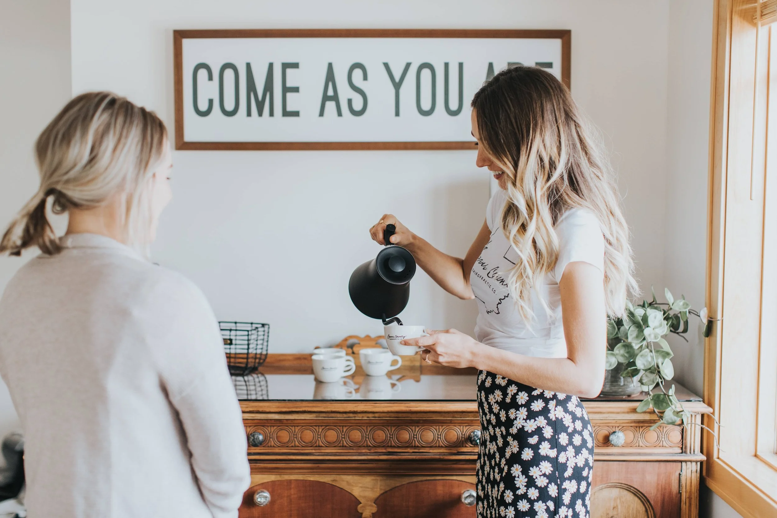 A woman smiling and holding a black handheld steam cleaner, serving a white mug to another woman in a cozy kitchen with wooden accents and a sign that reads 'Come As You Are'.
