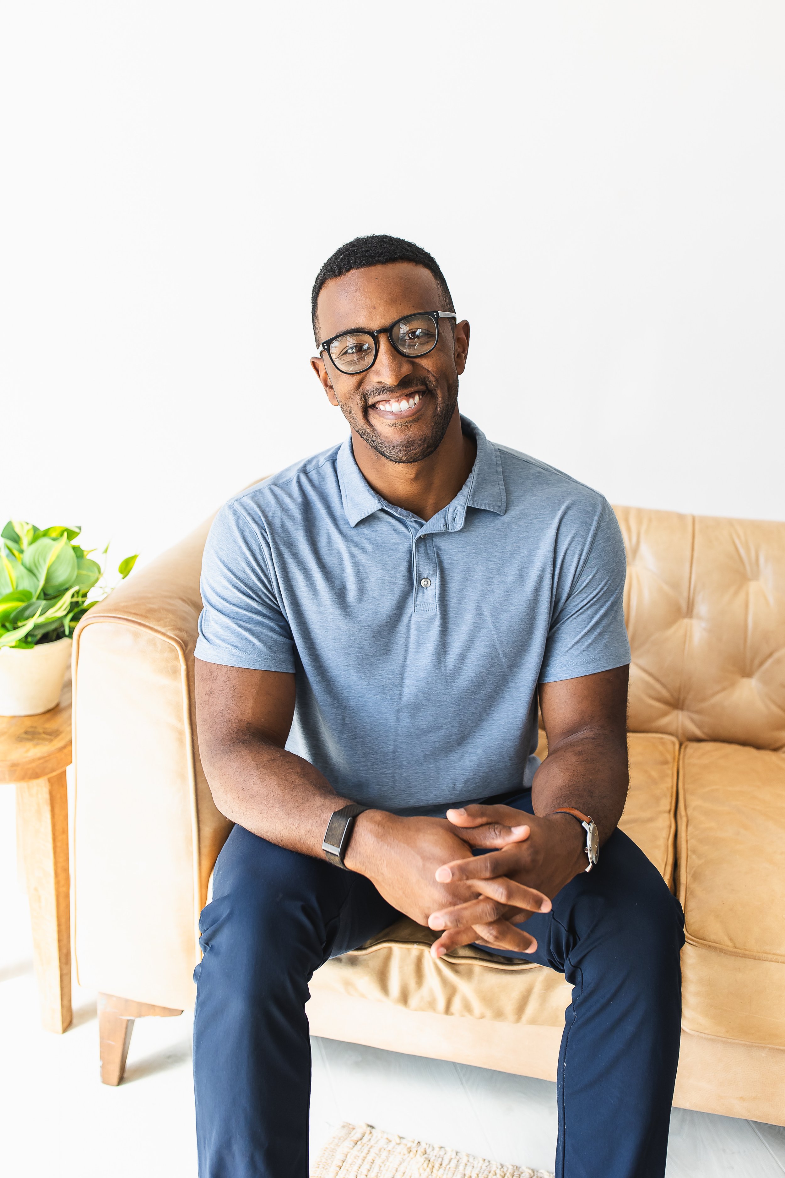 A man sitting on a beige couch with his hands clasped on his lap, smiling and wearing glasses, a blue polo shirt, black pants, and a wristwatch, with a green plant on a table beside him and a light-colored wall in the background.