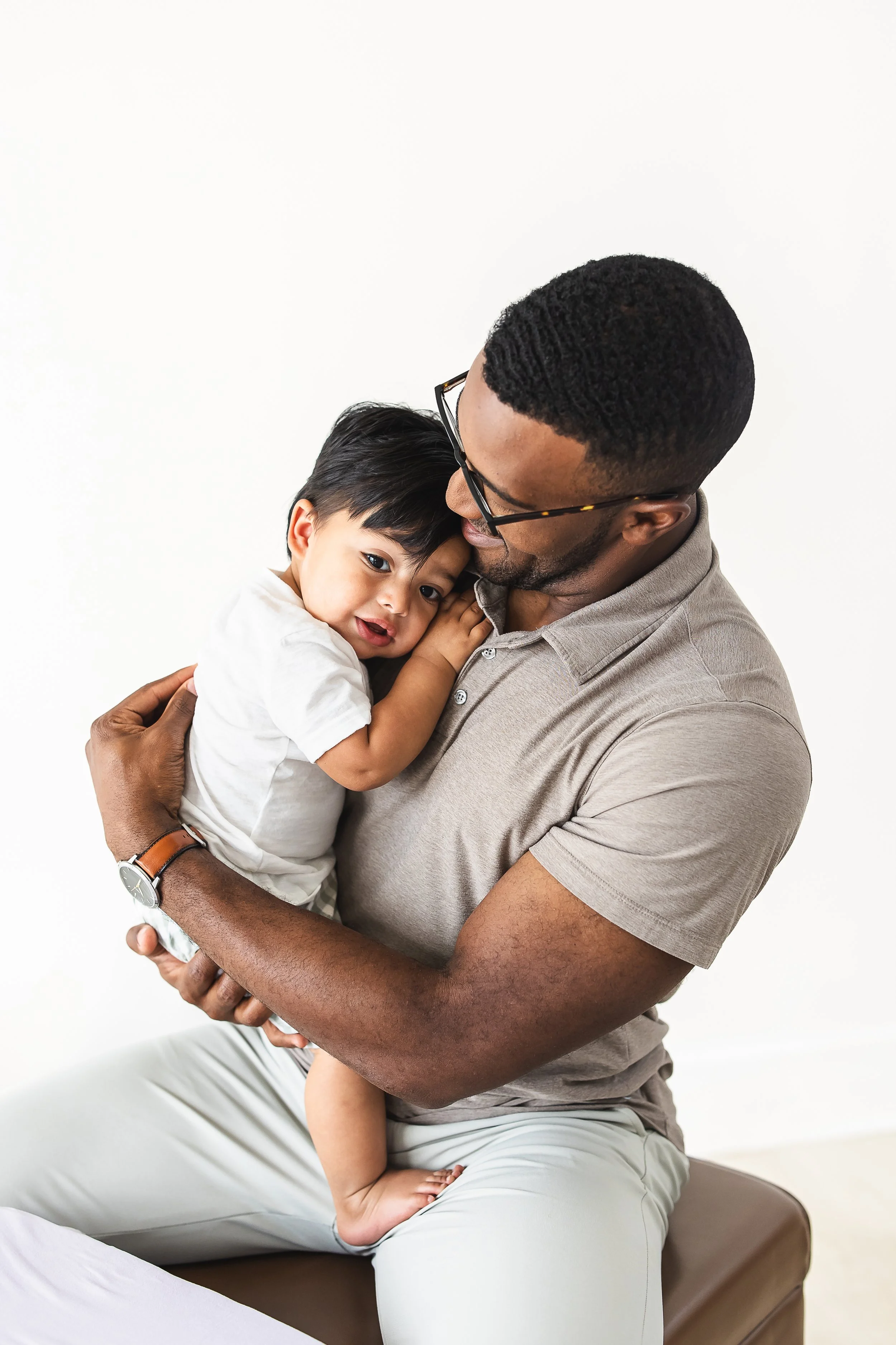 A man with glasses and a short beard holding a young child with short black hair and Asian features in his arms, both against a plain white background. The man is wearing a beige polo shirt and light-colored pants, and the child is wearing a white shirt and patterned shorts. They are sharing a close moment, with the child's face resting against the man's cheek.
