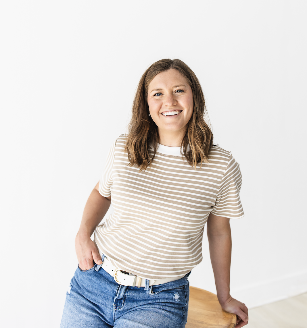 A woman with shoulder-length brown hair, wearing a beige and white striped t-shirt and blue jeans, smiling and leaning against a wooden surface in a bright, white room.
