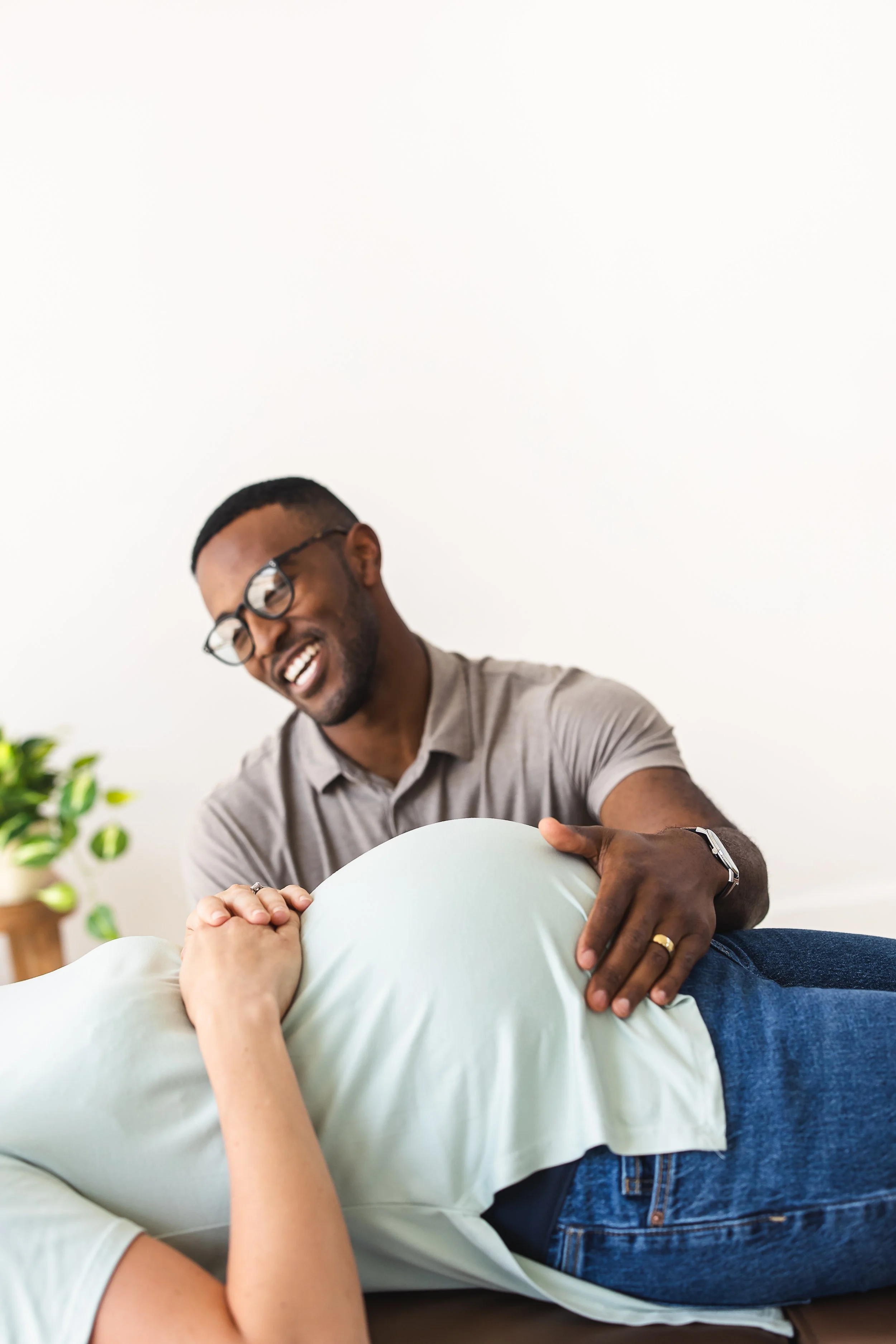 A smiling African American man with glasses and a beard, wearing a gray shirt, playfully pretending to give a massage to a woman lying on her stomach, dressed in a light green shirt, in a bright room with a white wall and a plant in the background.