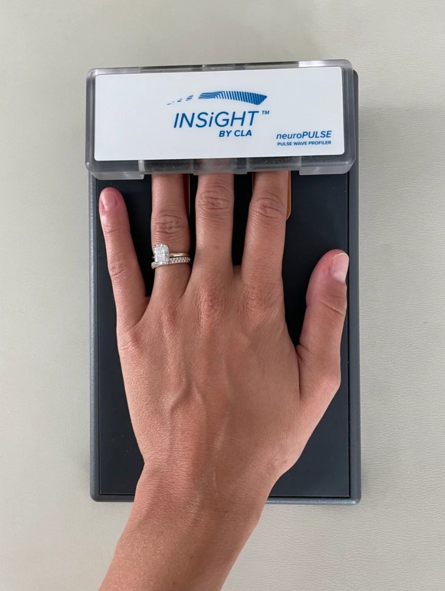 A person's hand resting on a black surface, with the ring finger adorned with rings, placed under a medical device labeled 'INSIGHT BY CLA neuroPULSE pulse wave profiler'.