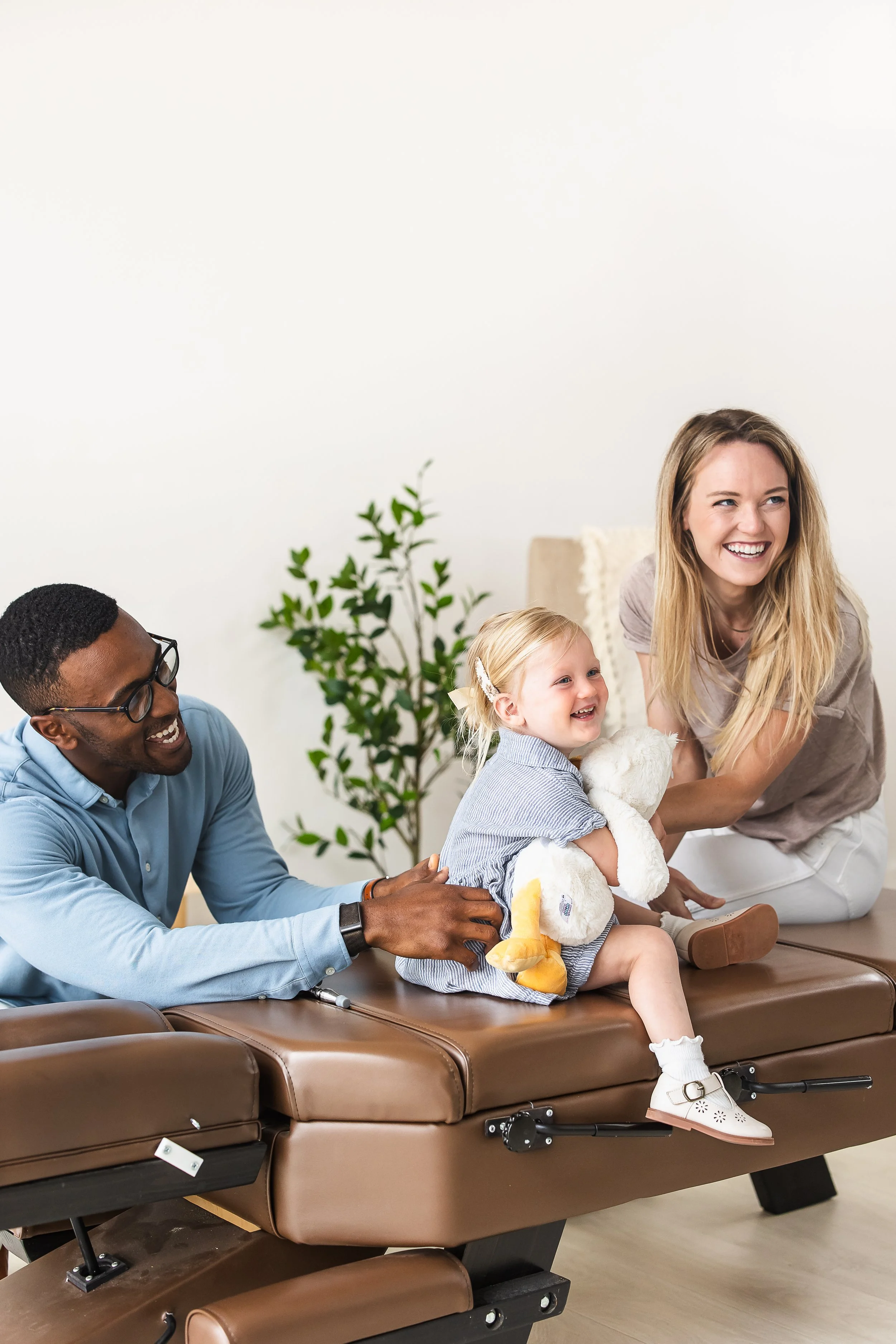 A group of people, including a young girl holding a stuffed animal, smiling and laughing, sitting around or near a brown therapy or examination bed in a bright, clean room with white walls and a potted plant.