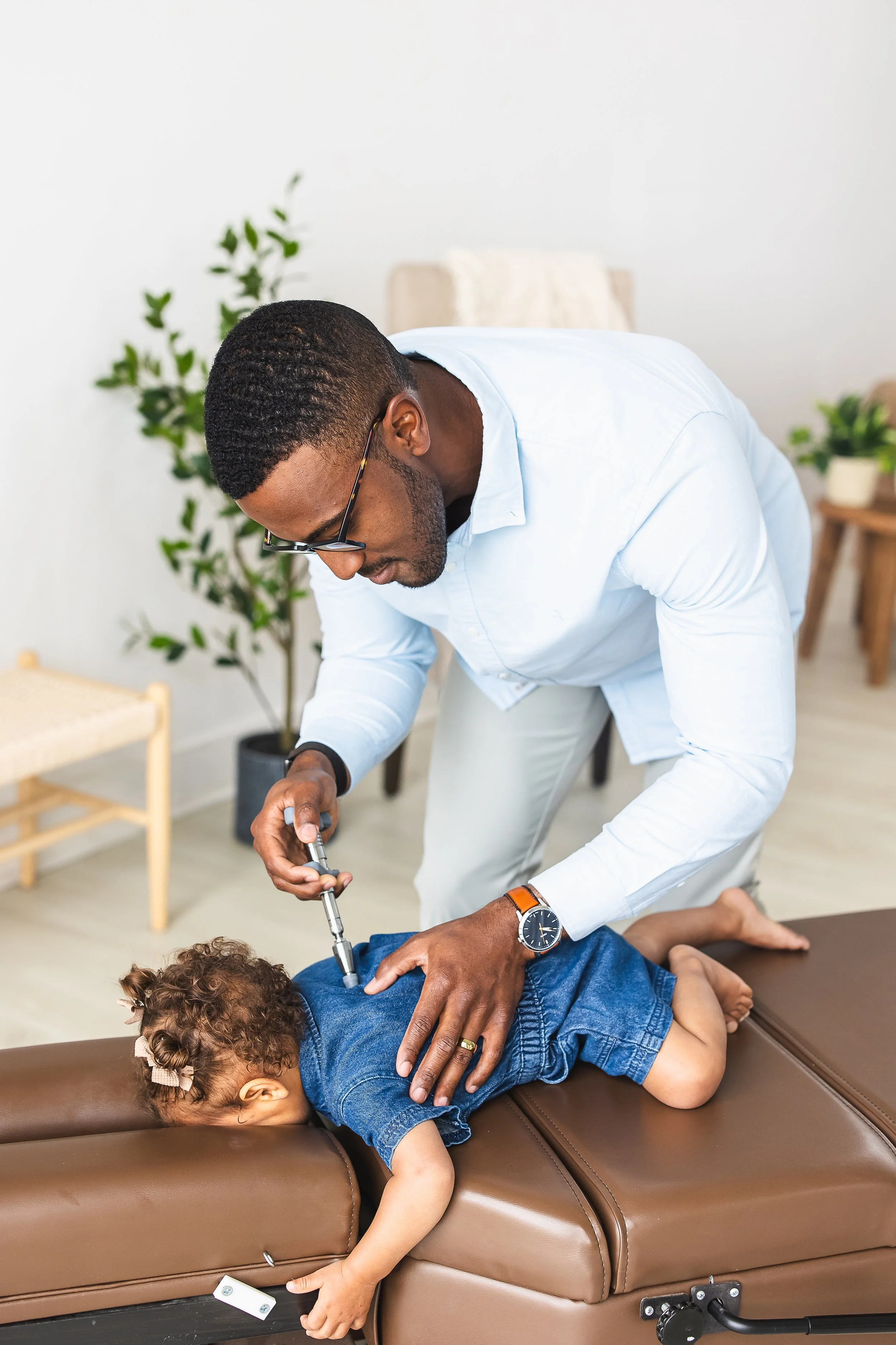 A chiropractor adjusting a young girl lying face down on a chiropractic table. The girl is wearing a blue dress and has a bow in her hair. The chiropractor is a man wearing glasses, a light blue shirt, and a wristwatch, using a tool on the girl. The room has a white wall, green plants, and a wooden table in the background.