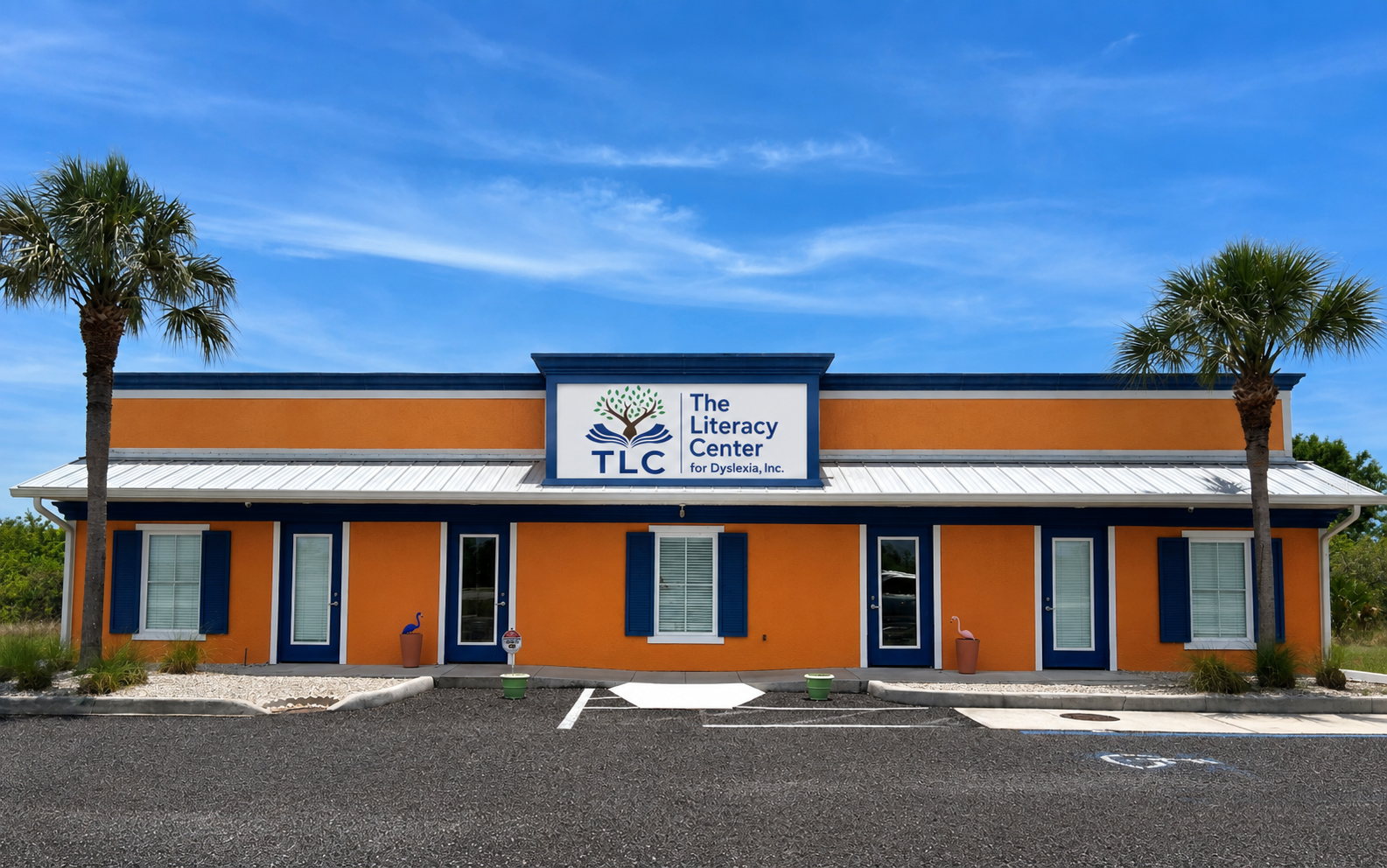 The Literacy Center building with orange walls, blue trim, and a white roof, featuring a sign with a tree logo and the text 'The Literacy Center for Dyslexia, Inc.', and two palm trees on either side of the building, under a blue sky.