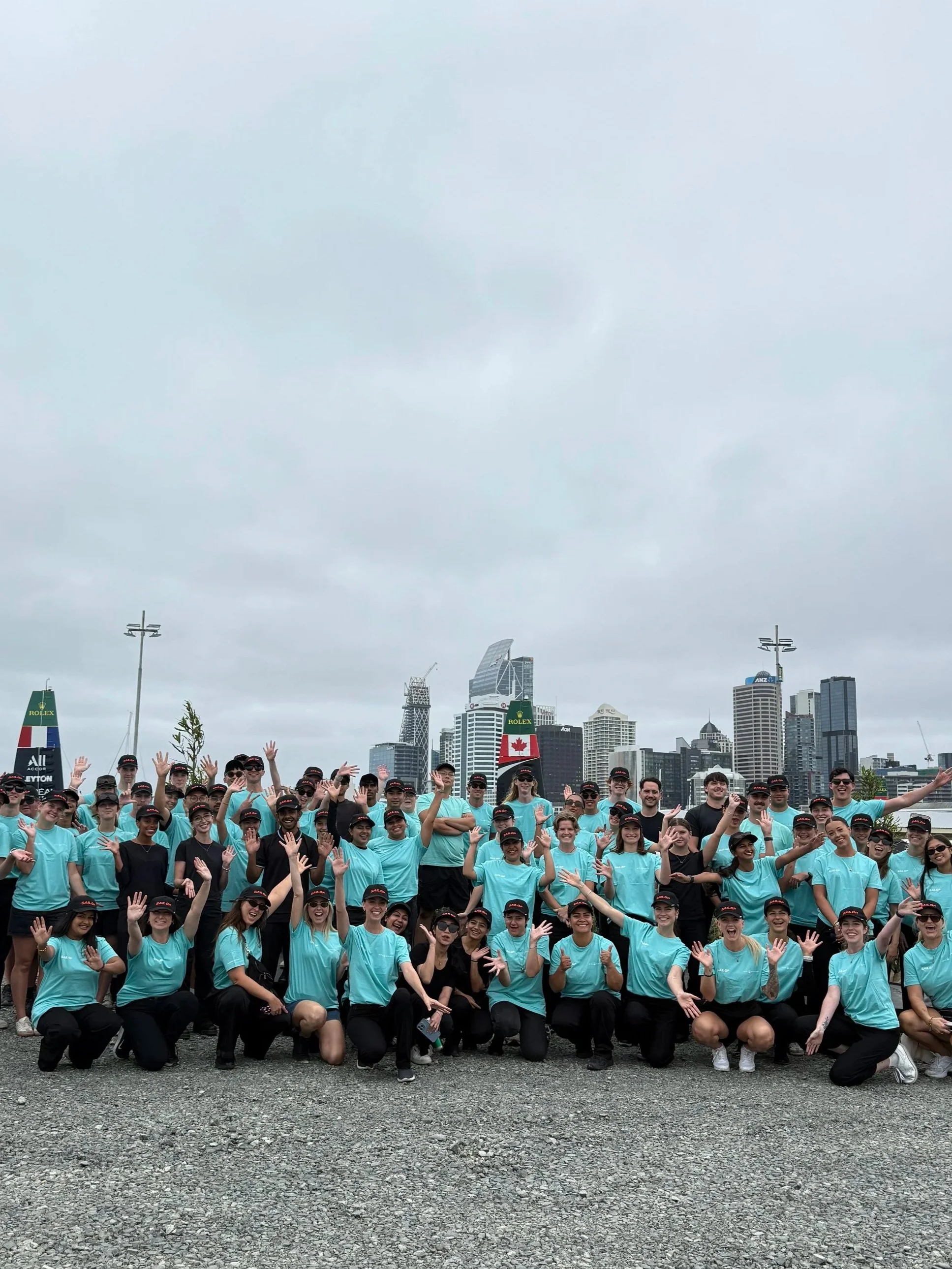 team of 70 staff dressed in blue sailgp tops on a cloudy auckland day celerbating with their hands up in a group photo