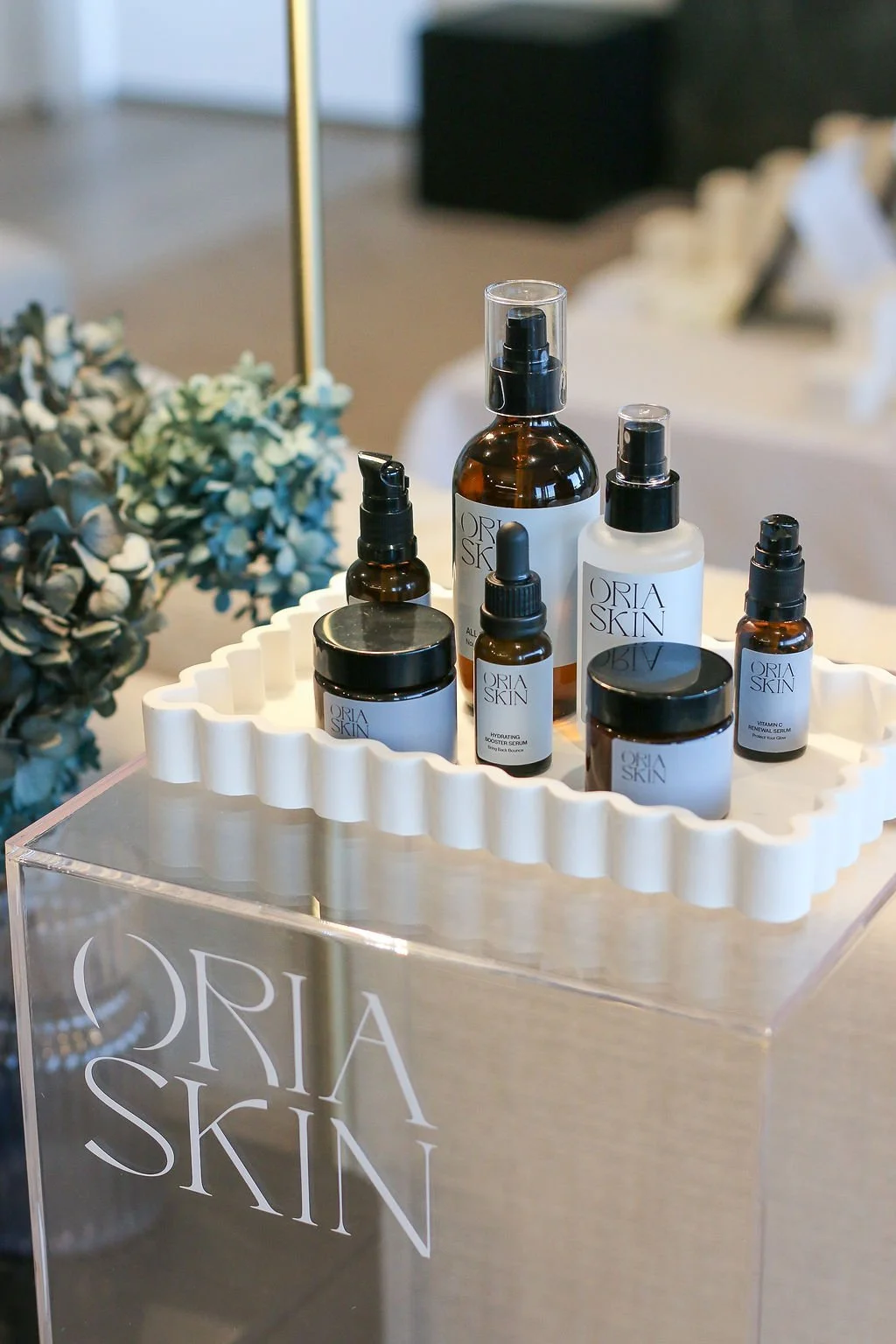 oria skin face product bottles on a white scallop edge tray in a clear plinth with oria skin decal. colours are muted soft cream whites and shades of blue