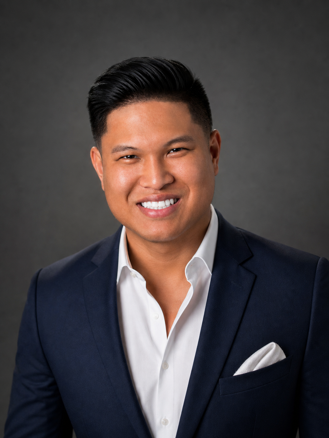 Professional headshot of a smiling man in a navy suit with a white shirt and pocket square, against a gray background.