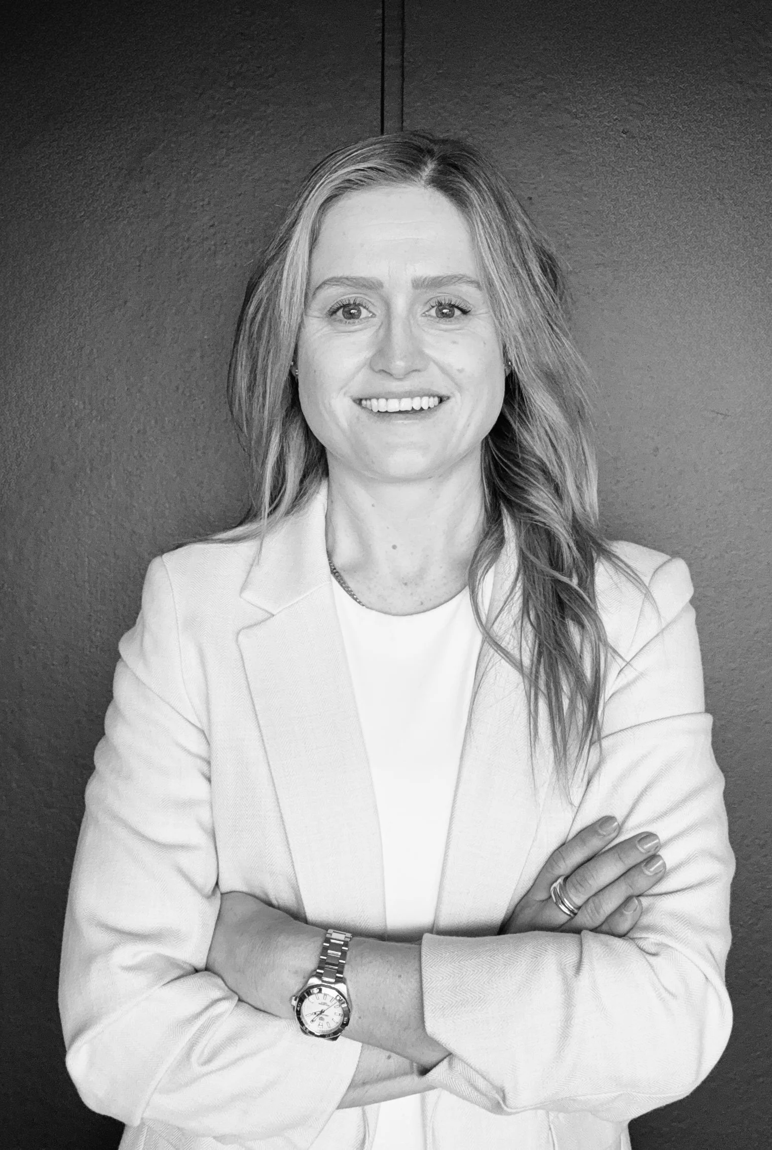 Black and white photo of a woman with light-colored hair, smiling, wearing a blazer and a watch, crossing her arms, against a plain dark background.