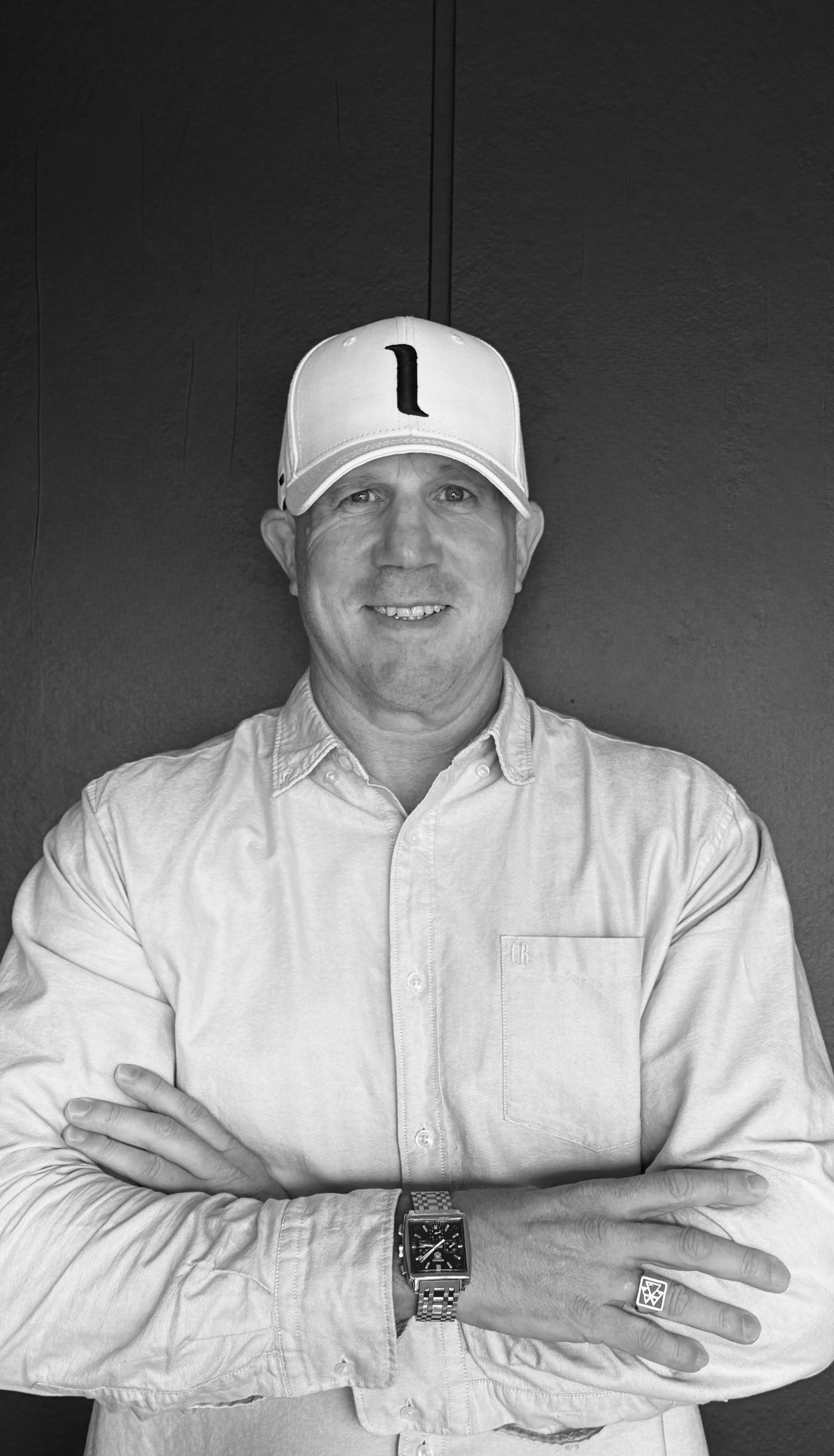 A man wearing a baseball cap with the letter 'I' on it, a button-up shirt, and a large watch on his left wrist, standing against a dark background with his arms crossed.