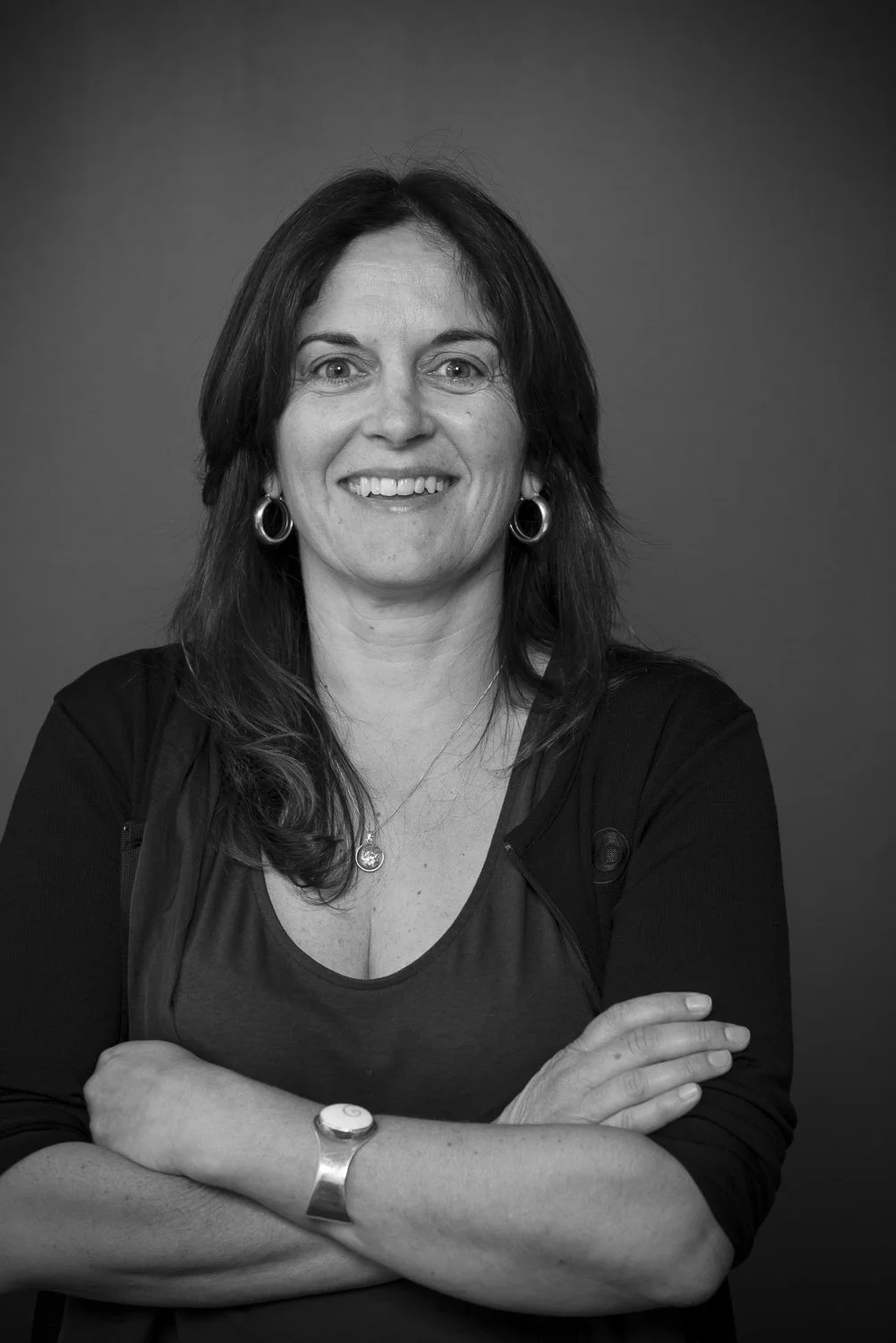 Black and white portrait of a woman with shoulder-length dark hair, smiling, wearing hoop earrings, a necklace, a watch, and a dark top with a jacket, posing with arms crossed against a plain background.