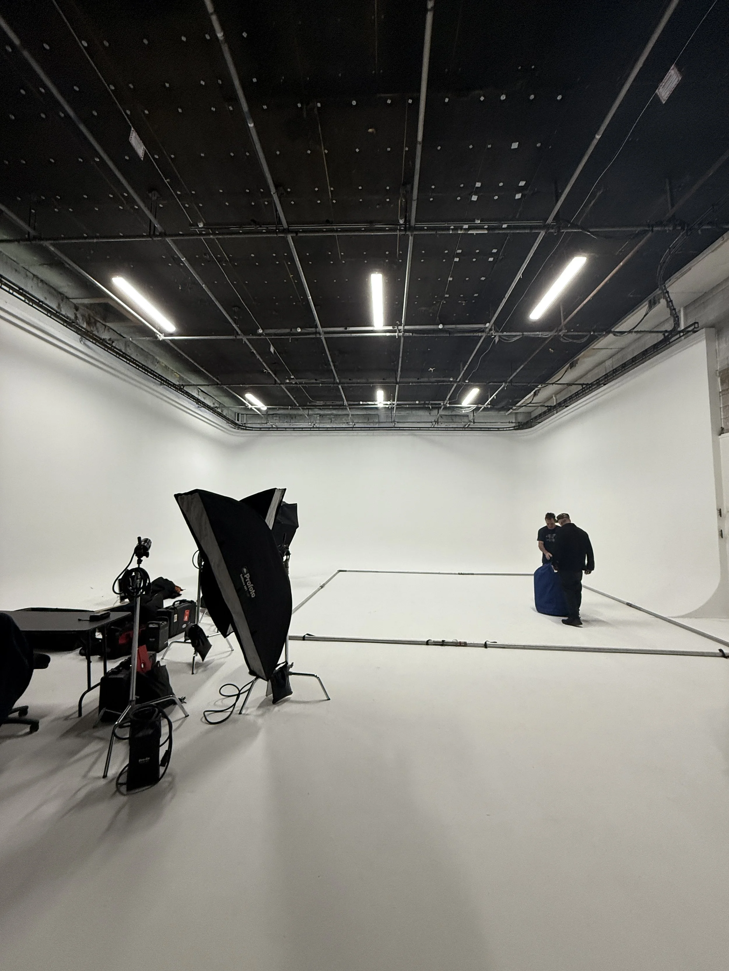 Photoss of a professional photography studio with seamless white background, large softbox lighting, and two people setting up a scene.