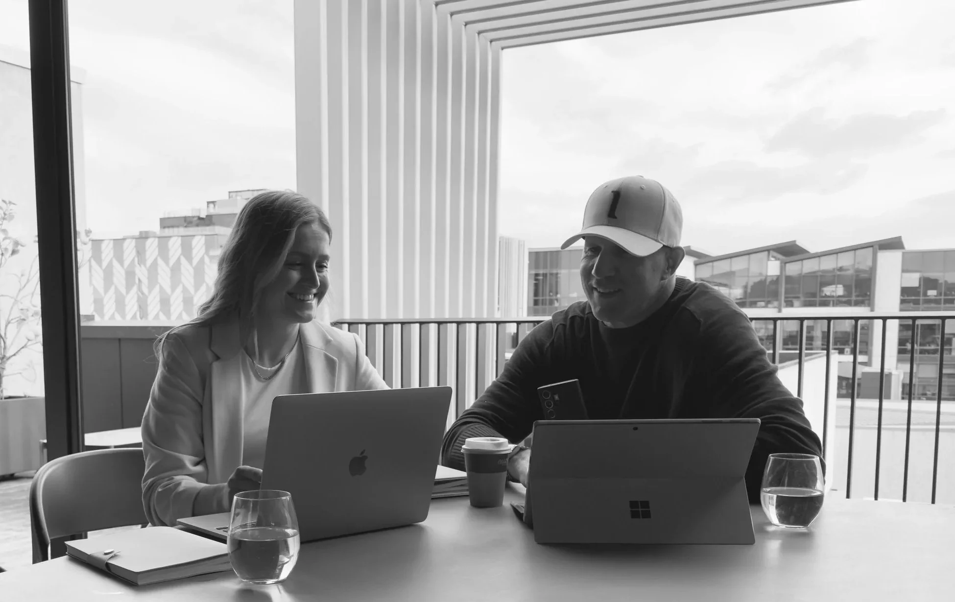 Two people sitting at a table with laptops, drinking coffee, smiling, and engaging in conversation inside a modern building with large windows showing an outdoor urban scene.