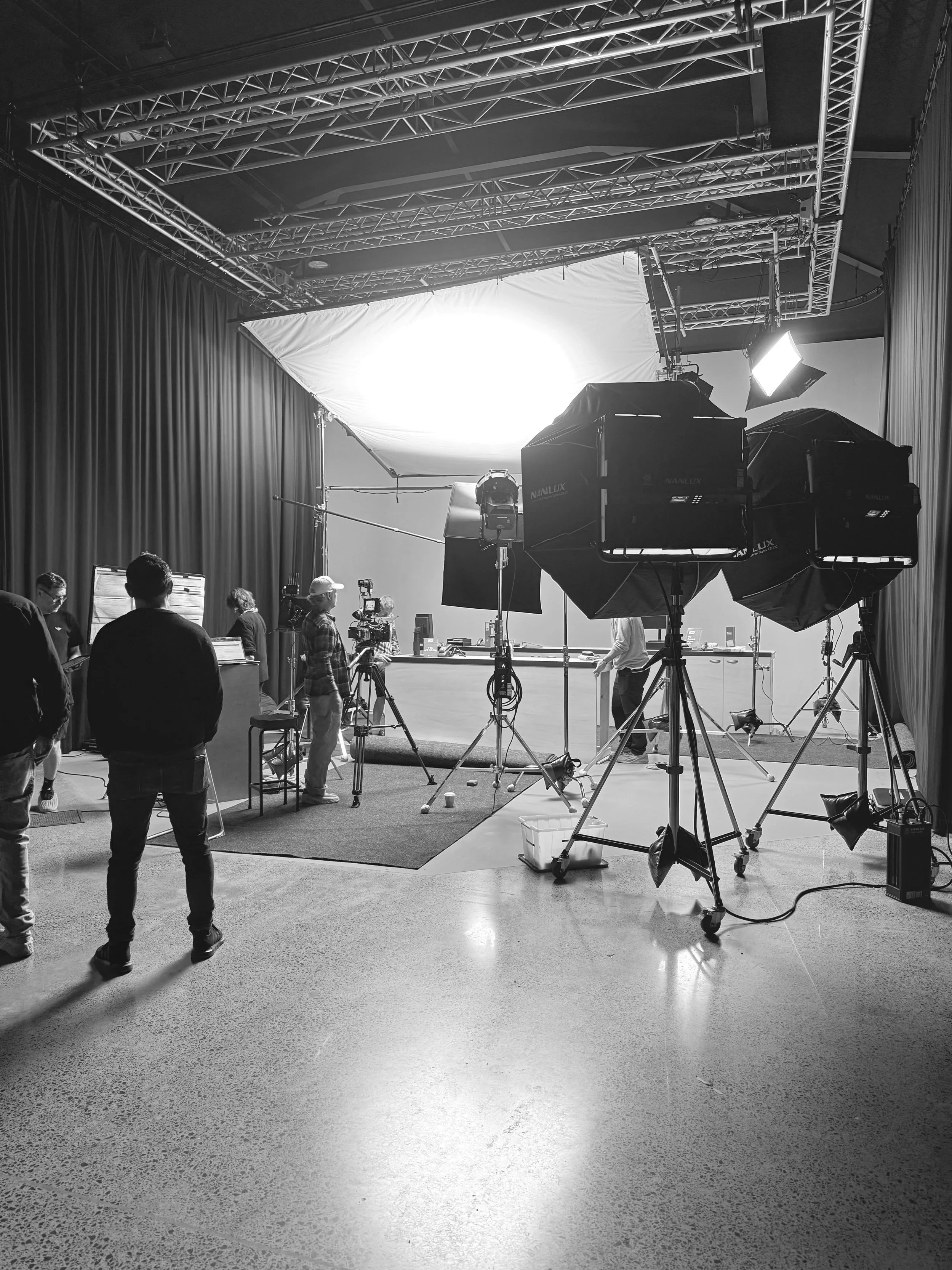 A black and white image of a film or photography studio with professional lighting equipment, curtains, and people working with cameras and computers.