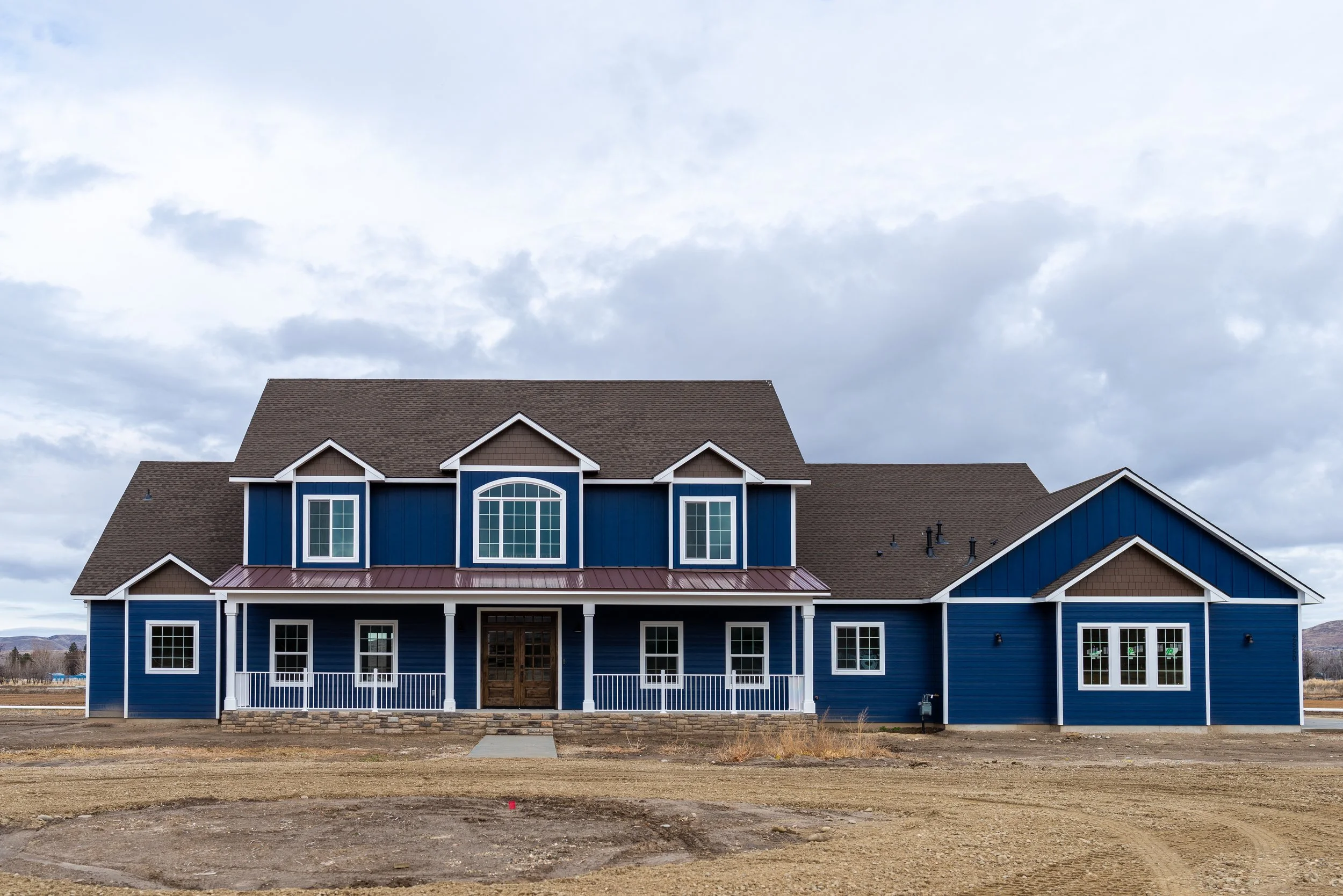Front view of a large two-story house with a brown roof, blue siding, multiple gabled windows, and a covered porch, set against a cloudy sky.