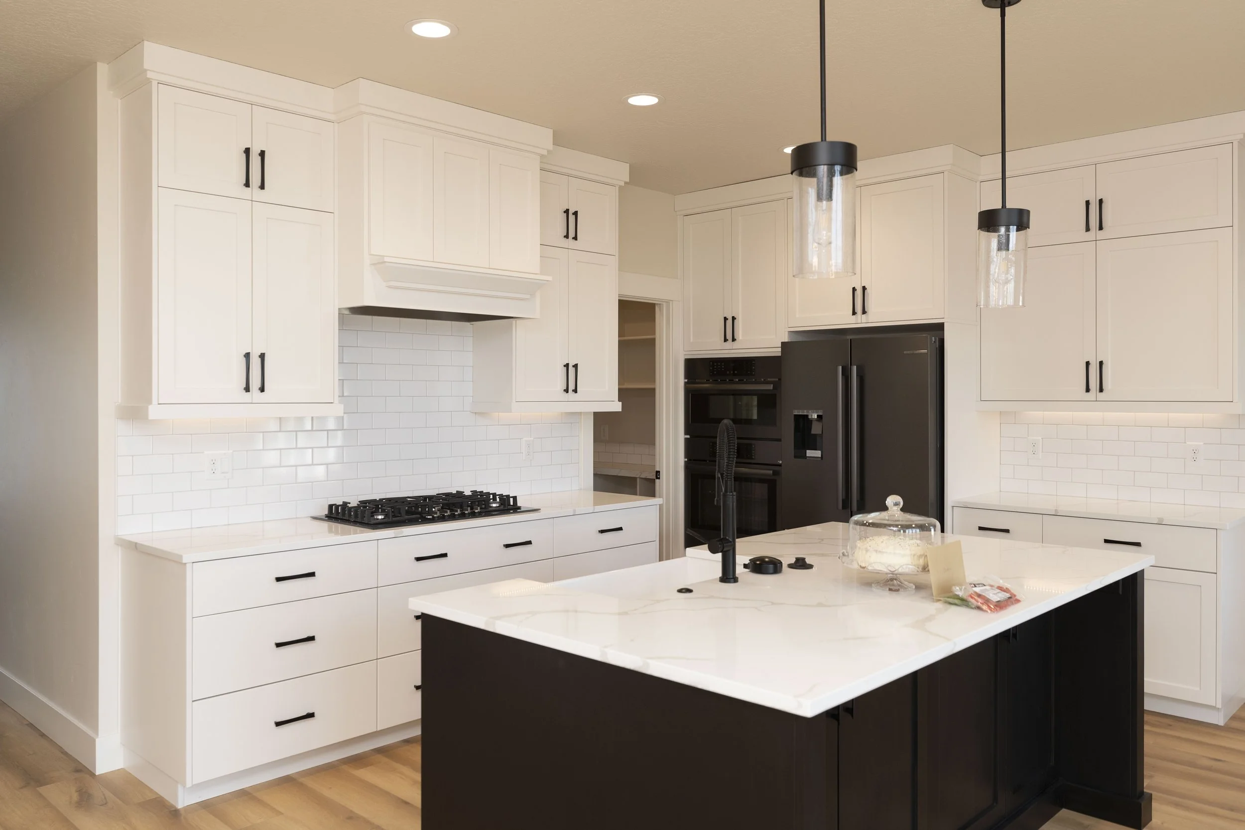 Modern kitchen with white cabinets, black hardware, a black and white island, and black pendant lights hanging over the island. Built-in black appliances include an oven and refrigerator. Wood flooring and a white subway tile backsplash complete the 