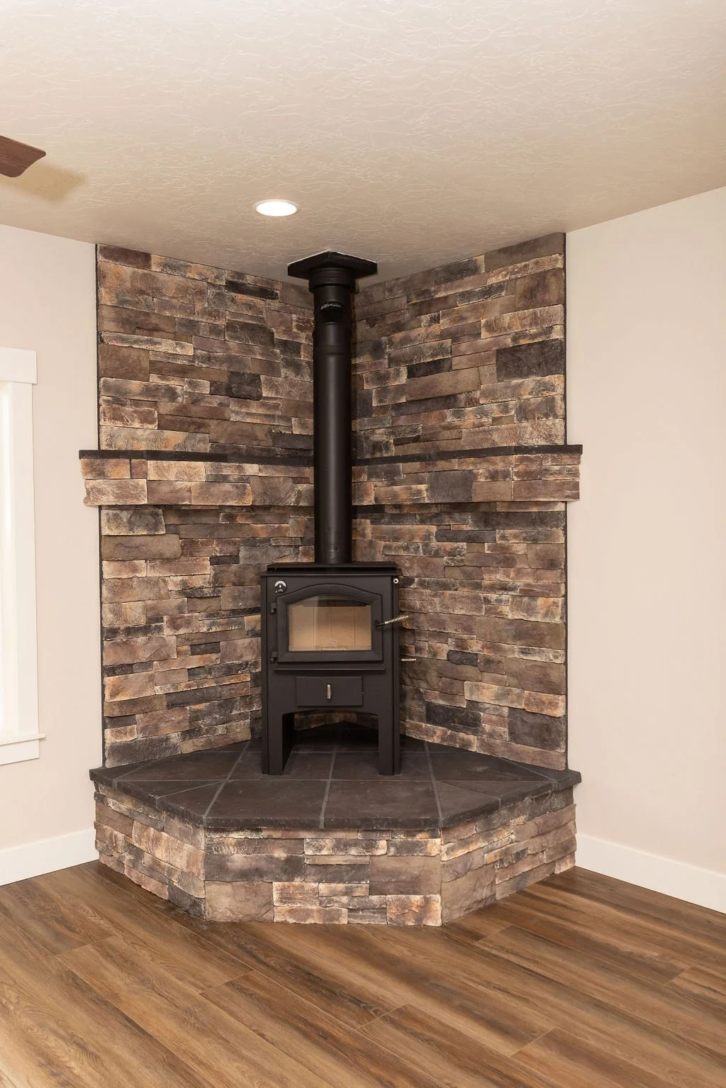 A corner fireplace with a black wood stove on a slate hearth, surrounded by a brown stacked stone wall, in a room with wood flooring and a white wall.