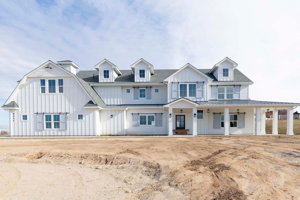 New white farmhouse-style house with gray roof and multiple windows under a partly cloudy sky.