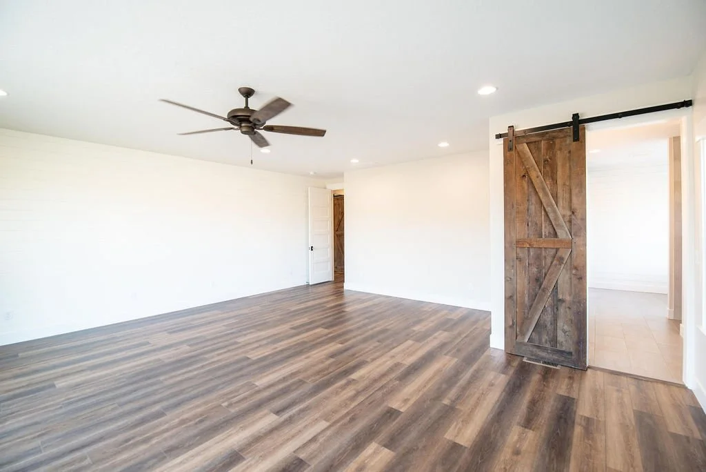 Empty living room with white walls, wood flooring, a ceiling fan, a sliding barn door, and a doorway leading to another room.