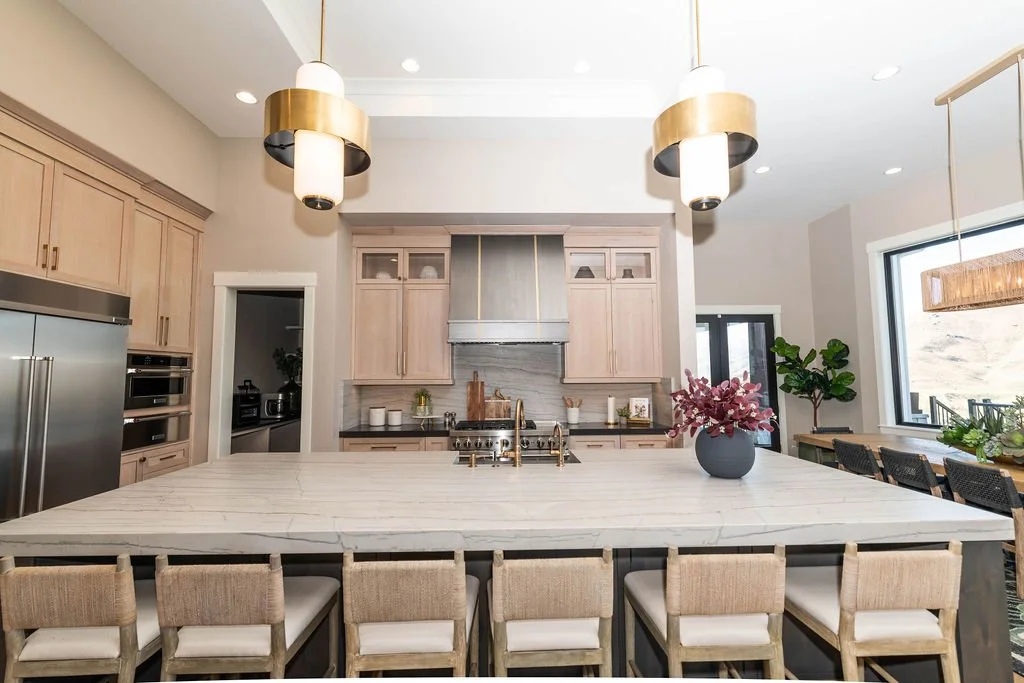 Modern kitchen with a large marble island, light wood cabinets, stainless steel appliances, two gold and white pendant lights above the island, a pink flower vase, and natural light from large windows.