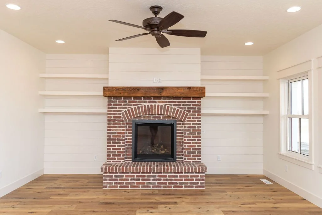 Living room with a brick fireplace, white shiplap walls, wooden mantle, three white shelves on each side, a ceiling fan, hardwood flooring, and a window on the right.