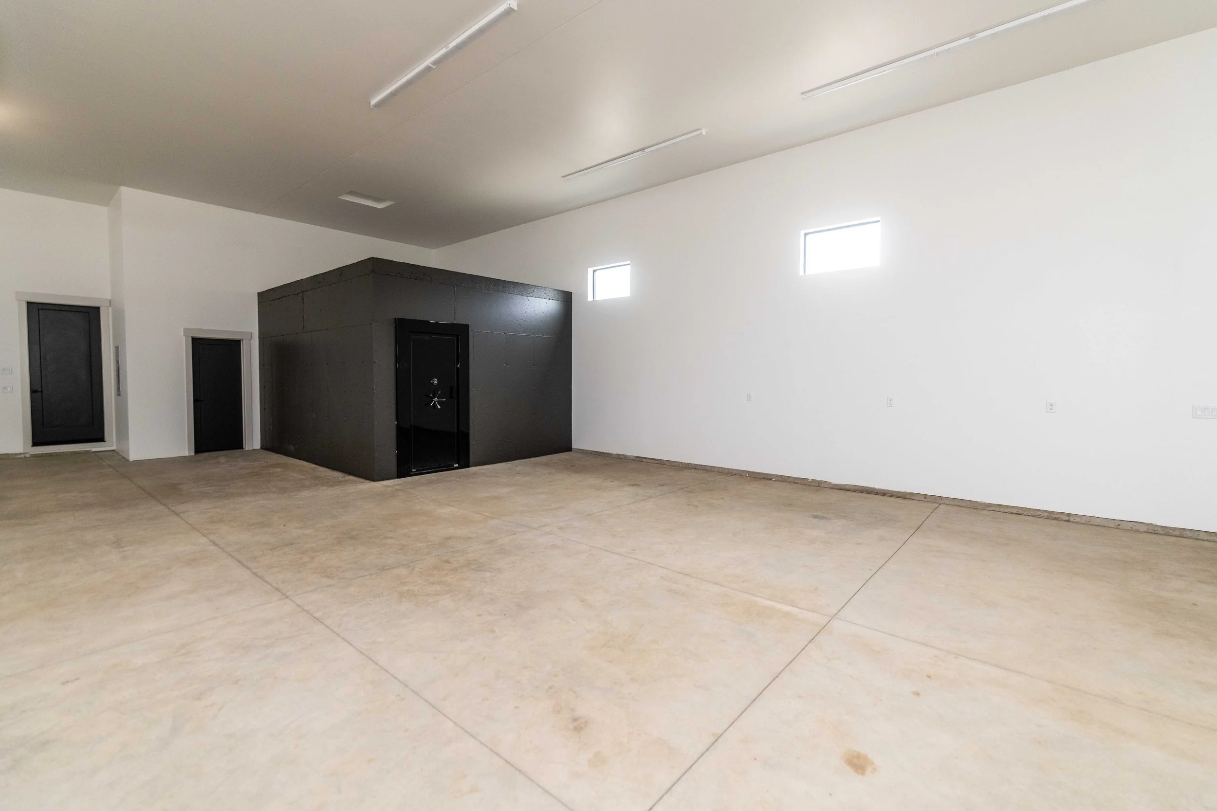 Interior of an empty room with white walls, concrete flooring, small rectangular windows near the ceiling, and a black vault-like structure with a door in one corner.