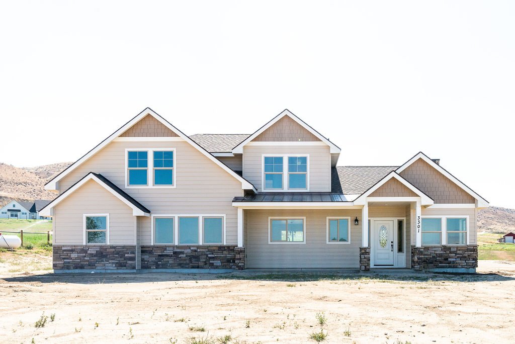Front view of a newly built, large suburban house with beige siding, stone accents, and multiple gabled roofs, located in a rural area with hills and open land.