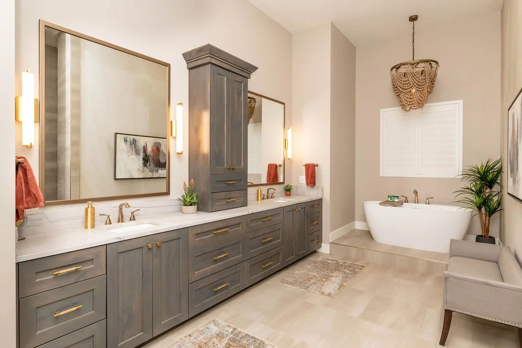 Bathroom with double vanity, large mirror, gold fixtures, and a bathtub on a raised platform. Decor includes potted plants, artwork, and wall-mounted lights.