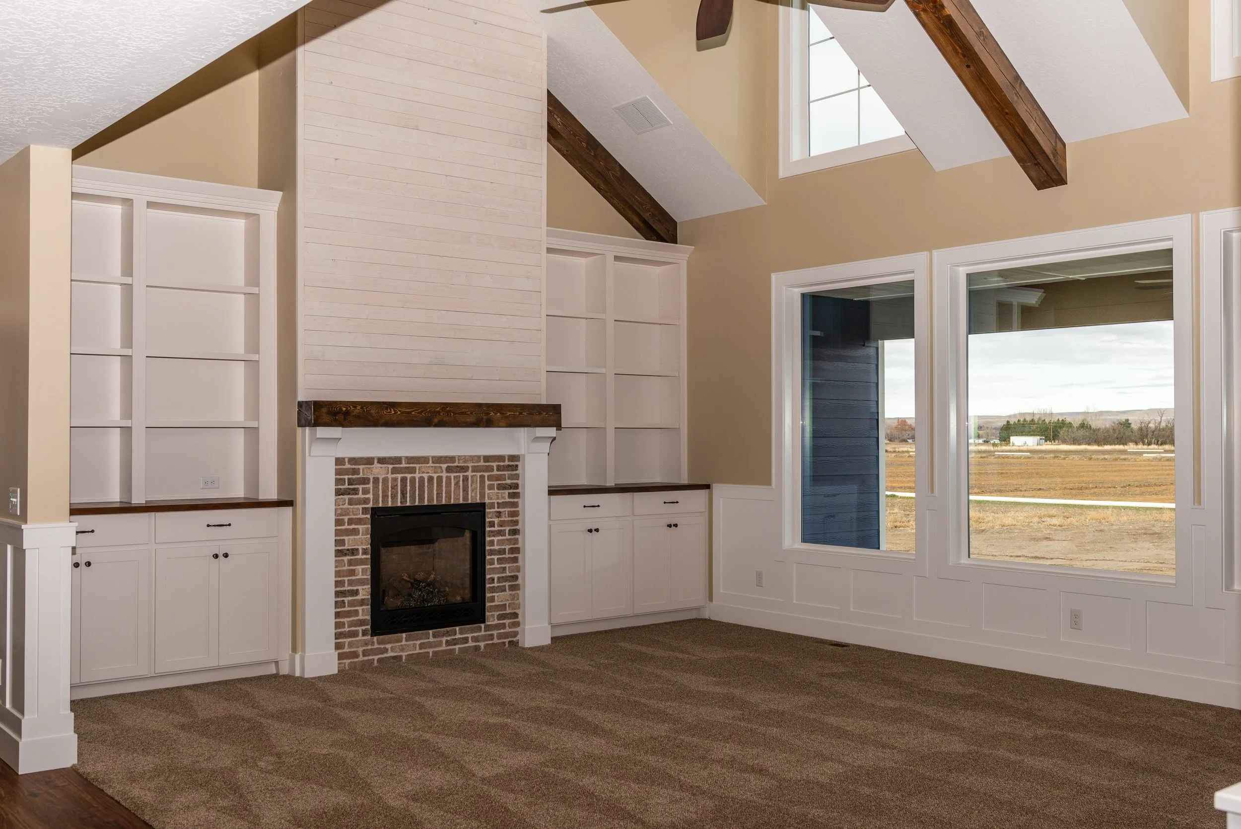Interior of a living room with a brick fireplace, white built-in shelves, large windows, and vaulted ceiling with dark wooden beams.