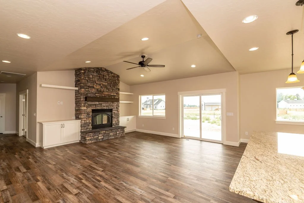 Spacious living room with a brick fireplace, large windows, and a sliding glass door leading outside, featuring wood flooring and a ceiling fan.