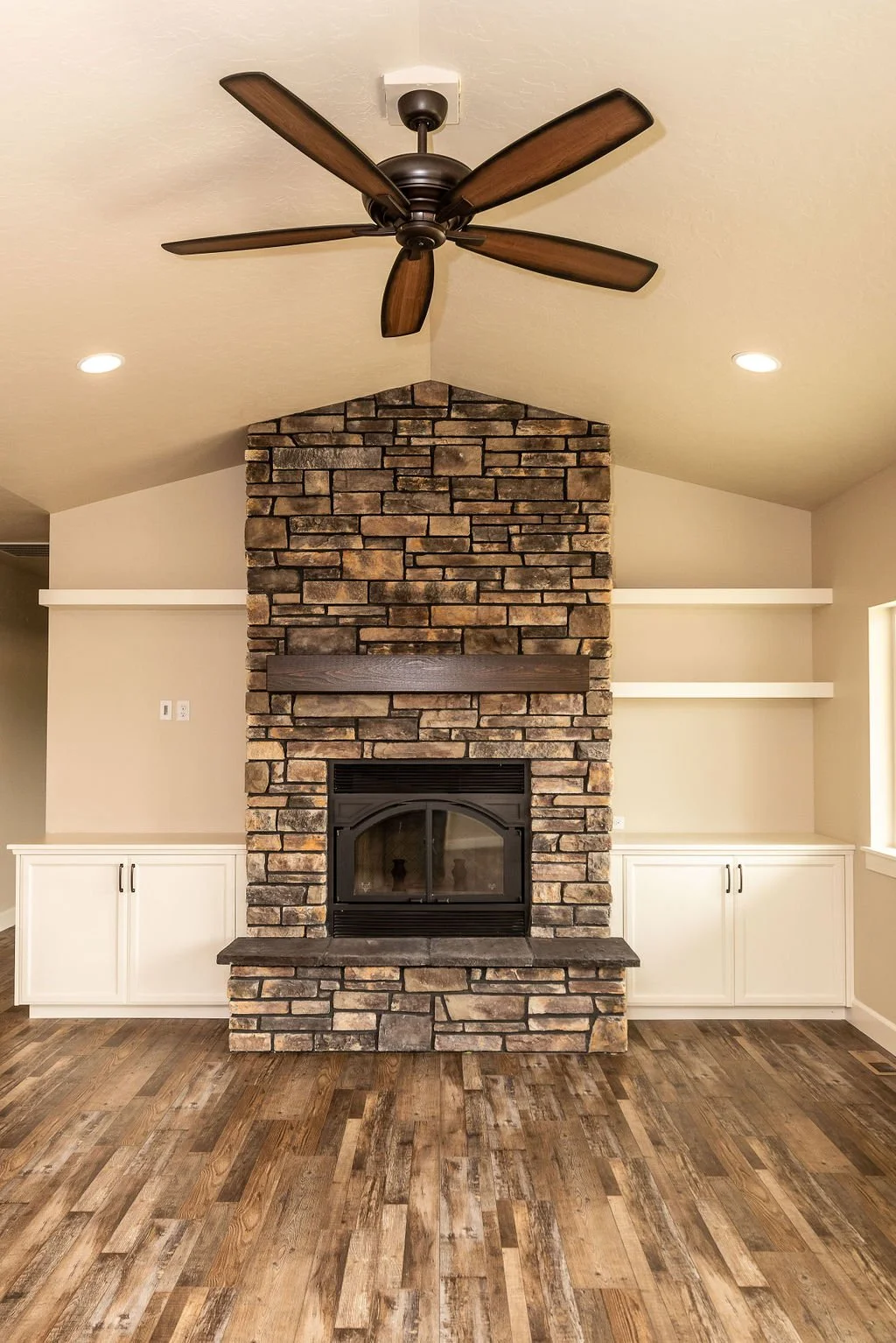 A living room with a stone fireplace, wooden mantel, white built-in cabinets, floating shelves, hardwood flooring, a ceiling fan, and recessed lighting.