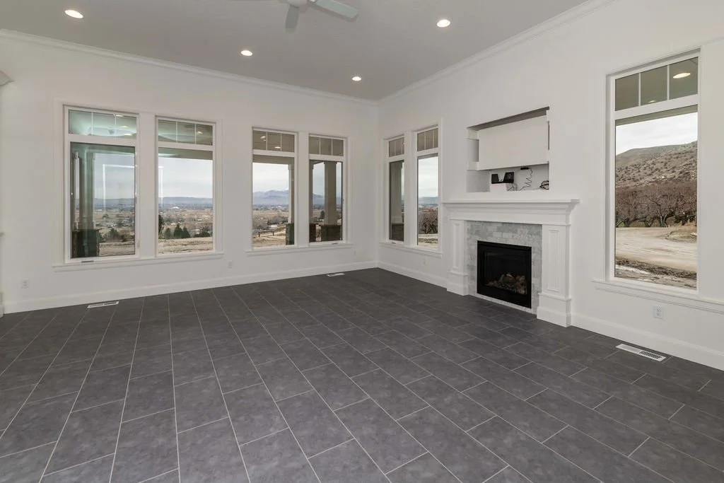 Empty living room with large windows overlooking a landscape, gray tiled floor, white walls, and a fireplace with built-in shelf above.