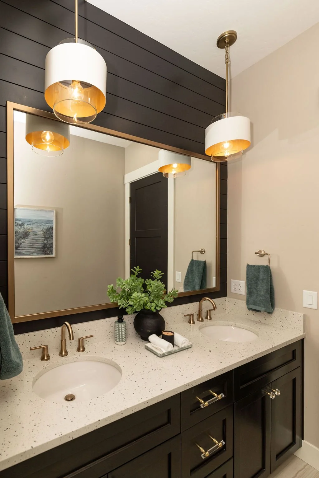 A modern bathroom double vanity with black cabinets, beige speckled countertop, two oval sinks, and gold fixtures. A large mirror with a gold frame is above the vanity, reflecting two hanging pendant lights and a dark door. Decorative items and green