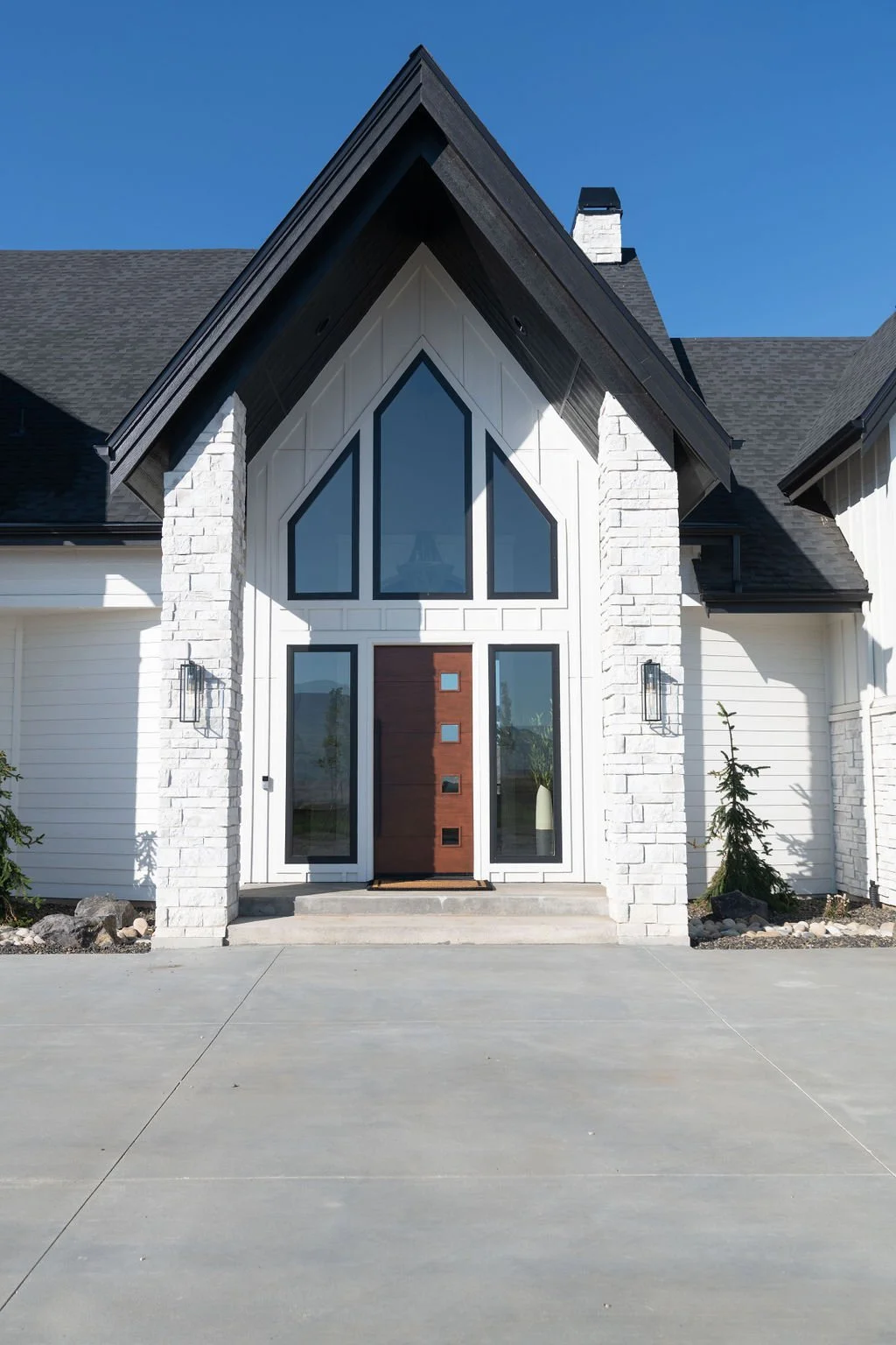 Front view of a modern house with white siding, brick accents, large triangular and rectangular windows, and a wooden front door, under a clear blue sky.