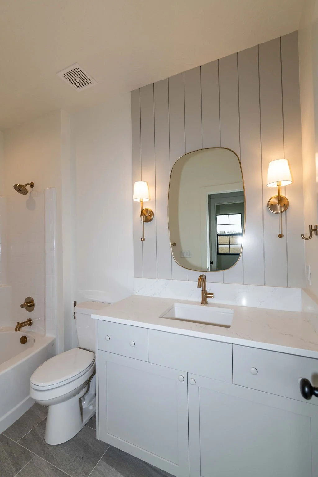 A modern bathroom with a white vanity, brass fixtures, a rectangular mirror, and wall-mounted lights.