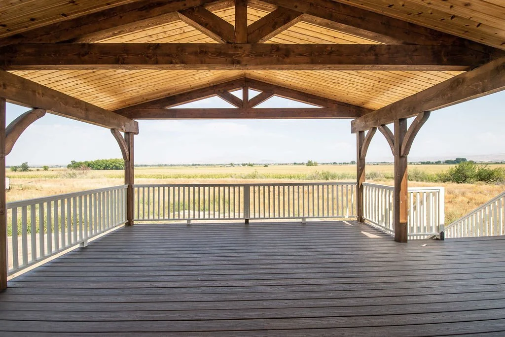 A spacious wooden outdoor deck with a railing, covered by a gabled roof with exposed wooden beams, overlooking a flat open field.