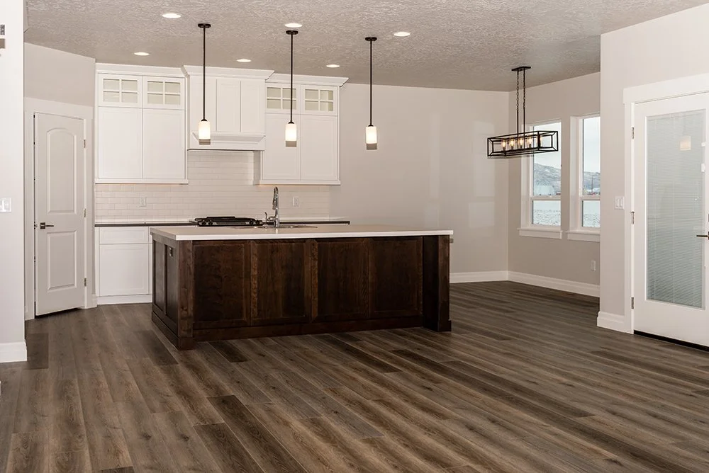 Empty modern kitchen with white cabinets, a white island with dark wood base, pendant lights, and wide plank wood flooring with a view of a water body through windows.
