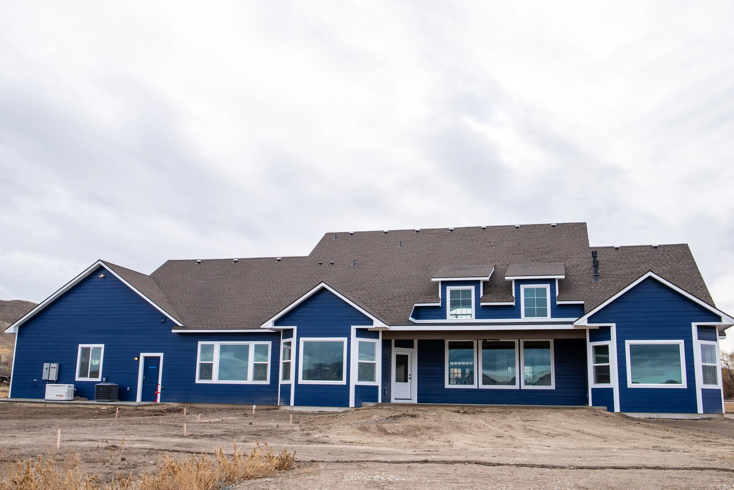 New blue house under construction with a brown shingle roof, large windows, and a front porch, set against a cloudy sky.